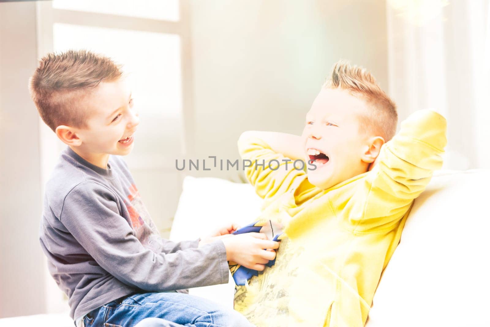 Two laughing young brothers tickling each other on a sofa as they relax having fun in a high key portrait
