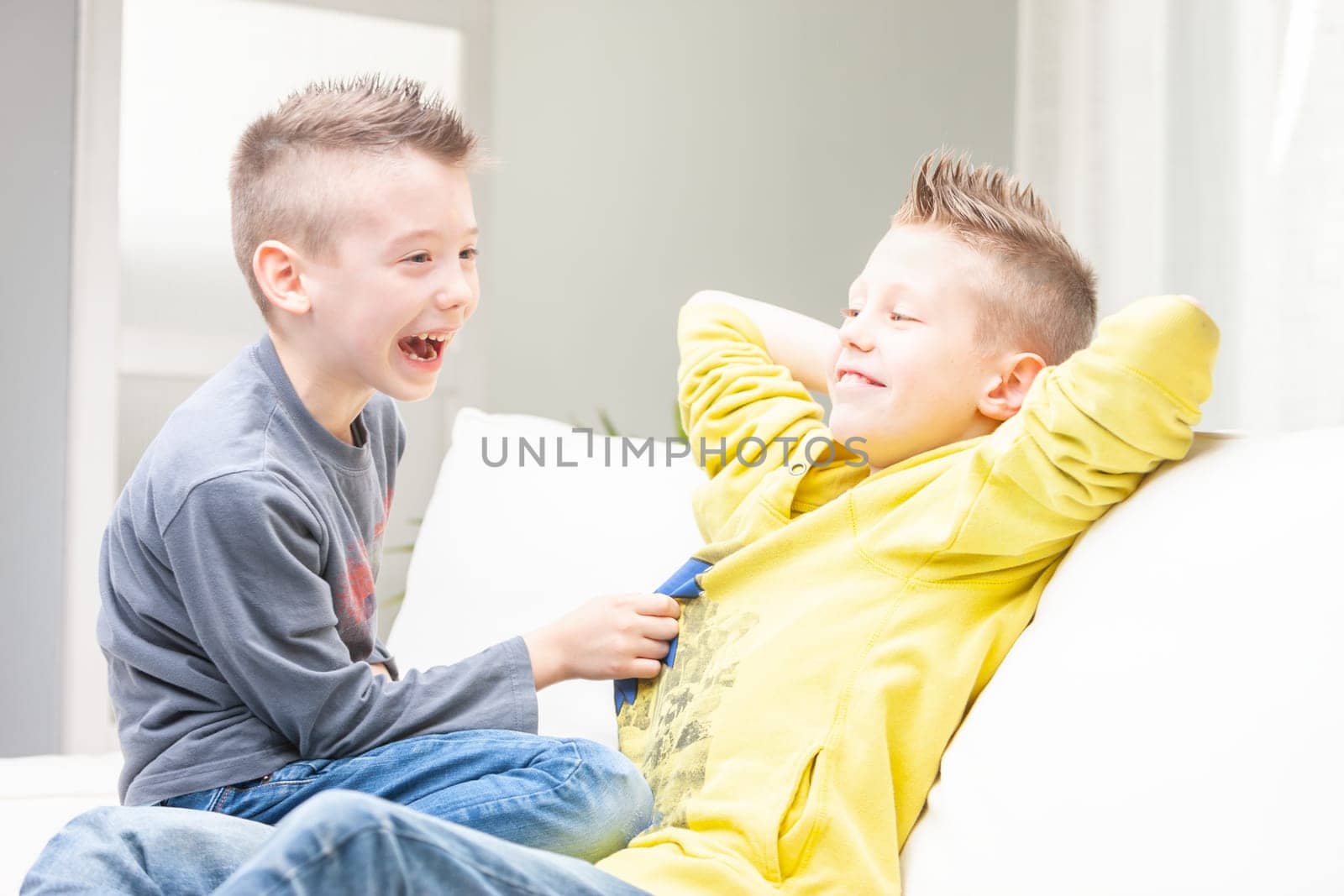 Cute funny young boy laughing at his brother as he tries to tickle him while they relax on the couch together at home