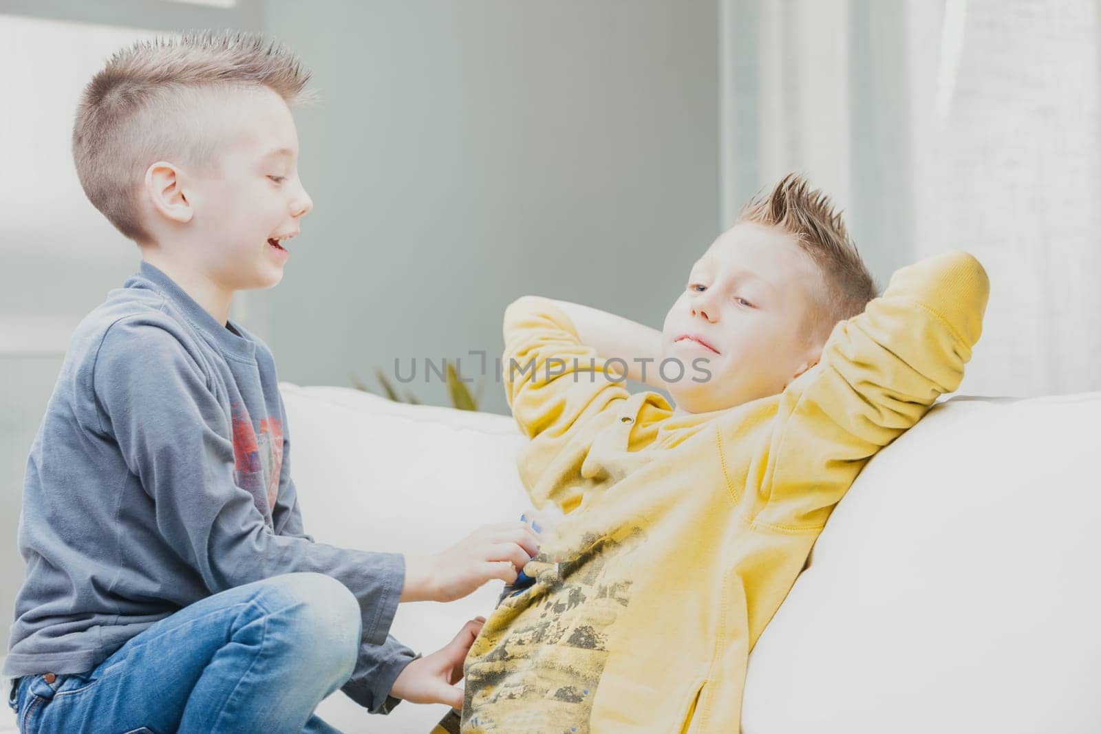 Mischievous young boy tickling his older brother on the stomach as they relax together on a sofa in a high key image
