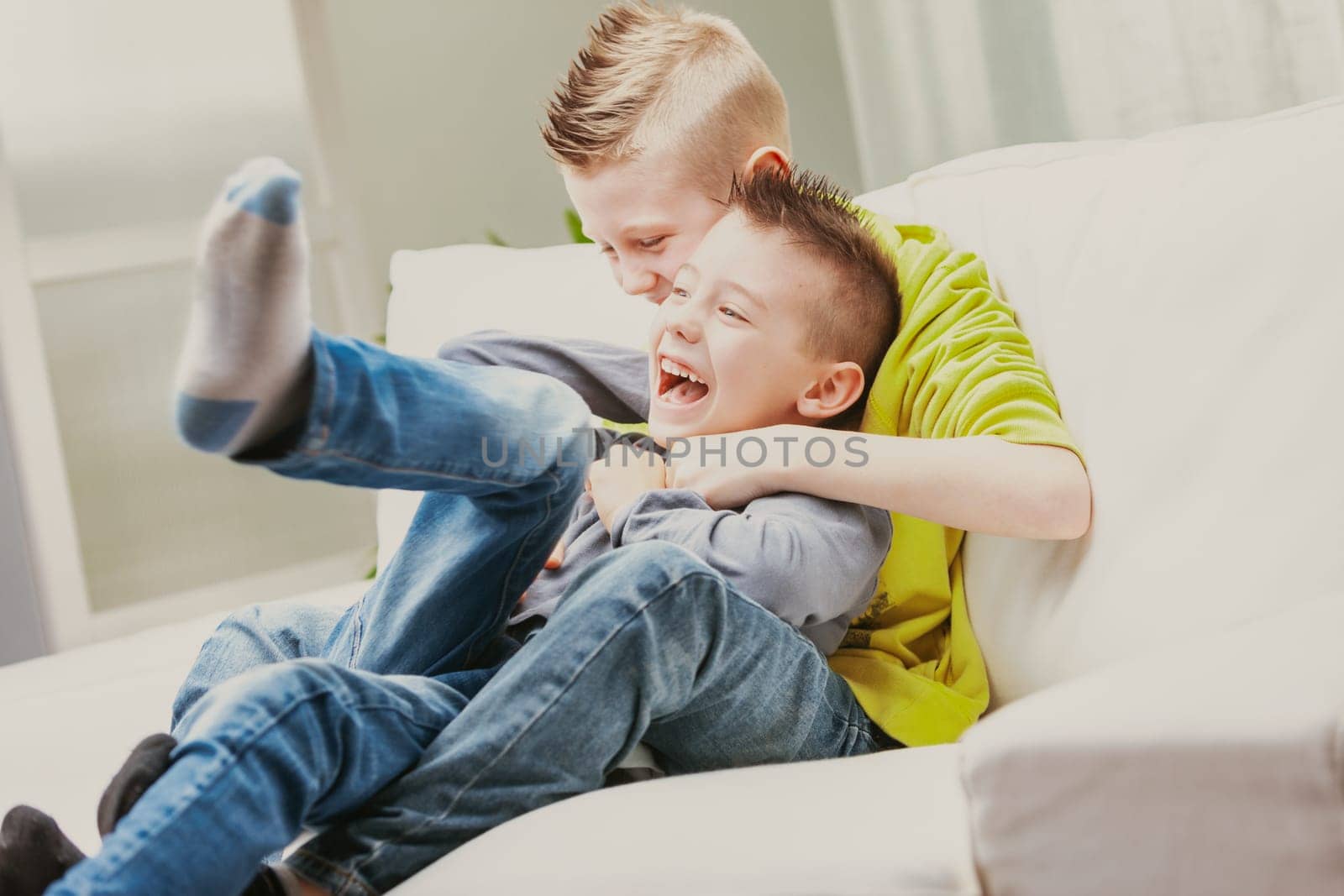 Two mischievous young brothers laughing as they romp on the couch in the living room in a high key candid portrait