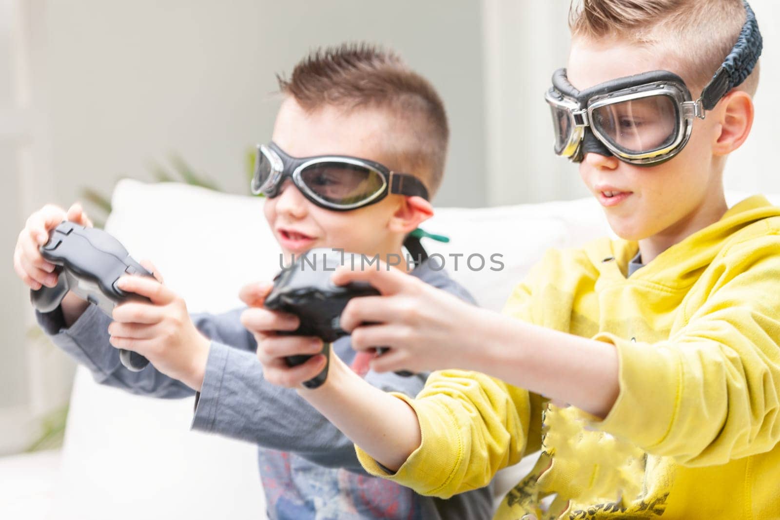 Two competitive young boys playing computer games seated side by side on a sofa with raised games controllers wearing goggles