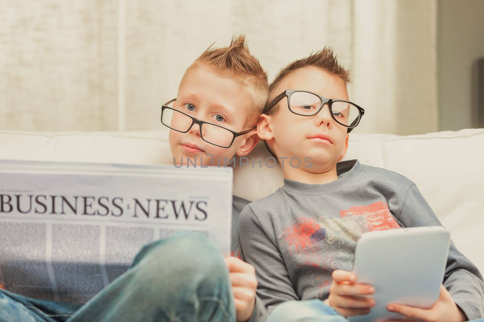 Two studious young brothers wearing glasses relaxing on a sofa, one with the Business Times and one with a tablet computer