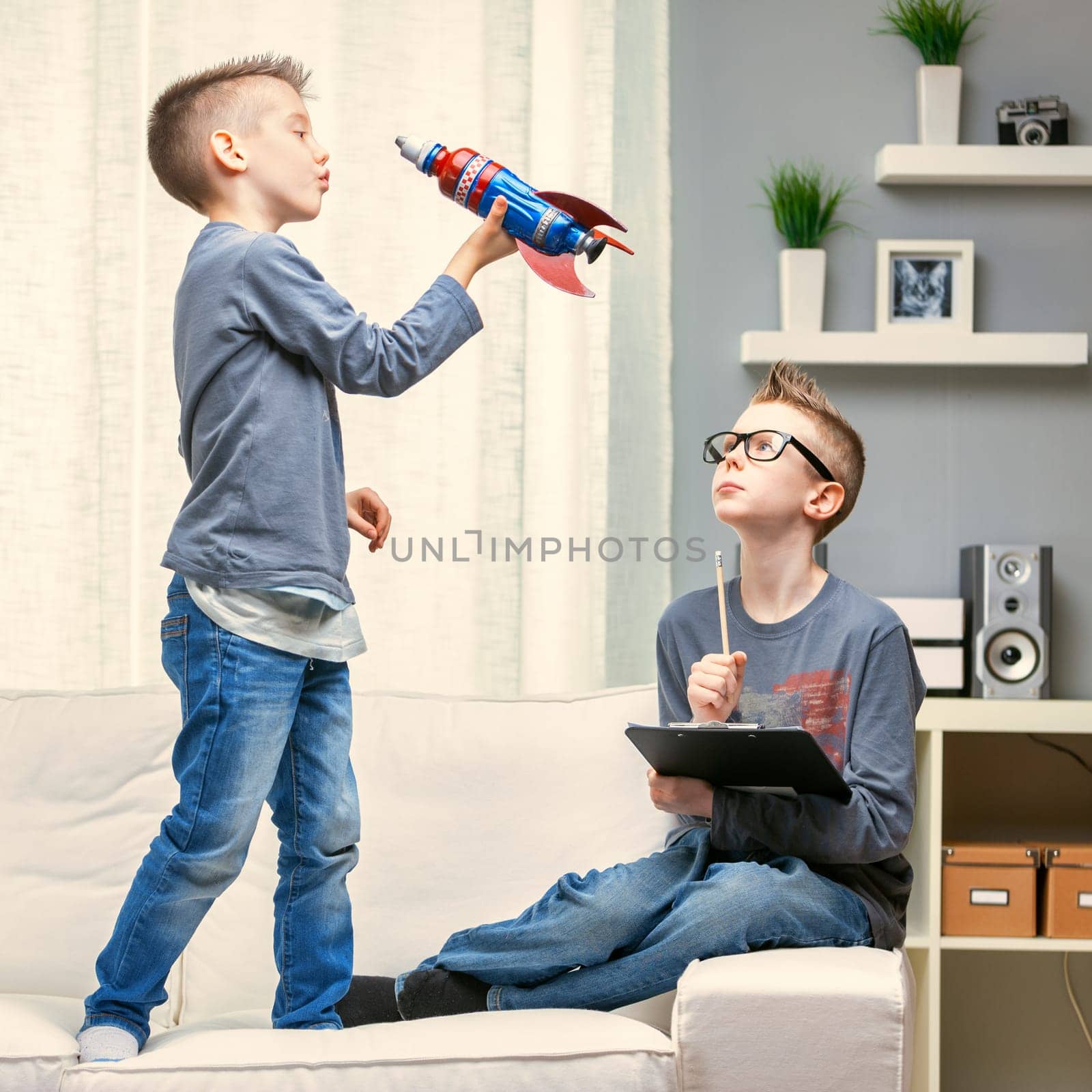 Cute young boy playing with a toy rocket on a sofa watched by his brother sitting alongside working on a clipboard