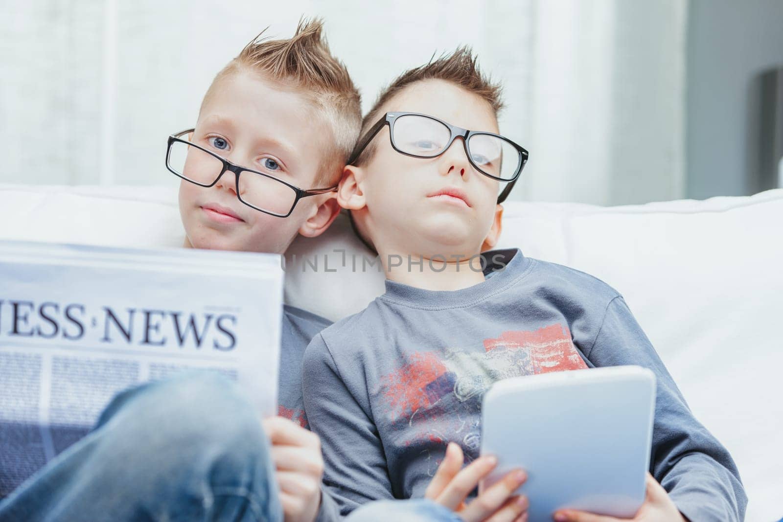 Portrait of two cute young boys in glasses sat at home with tablet and business newspaper
