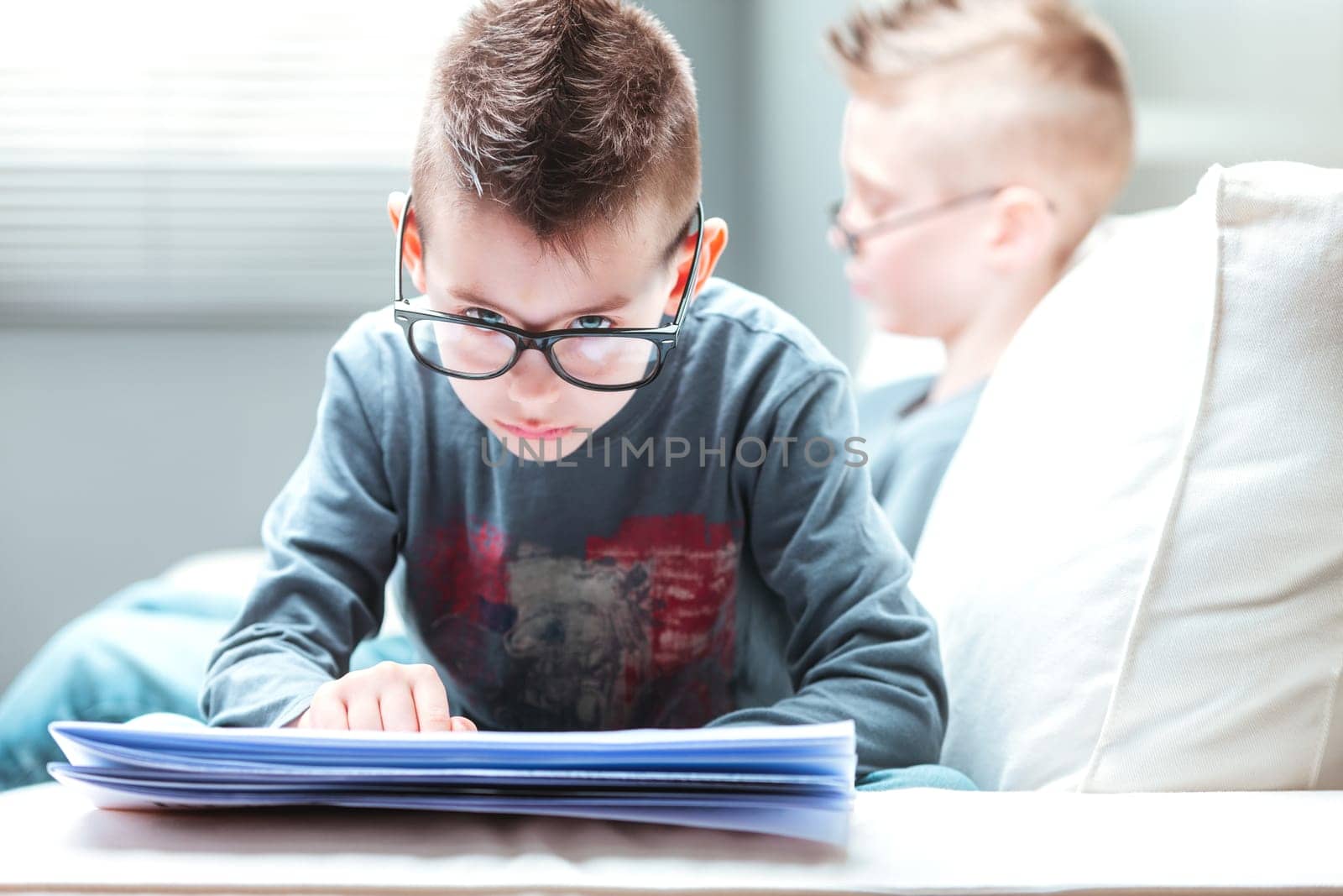 Young blue-eyed boy with upswept hairstyle relaxing reading a newspaper on the sofa with his brother or a friend as he peers over his glasses at the camera