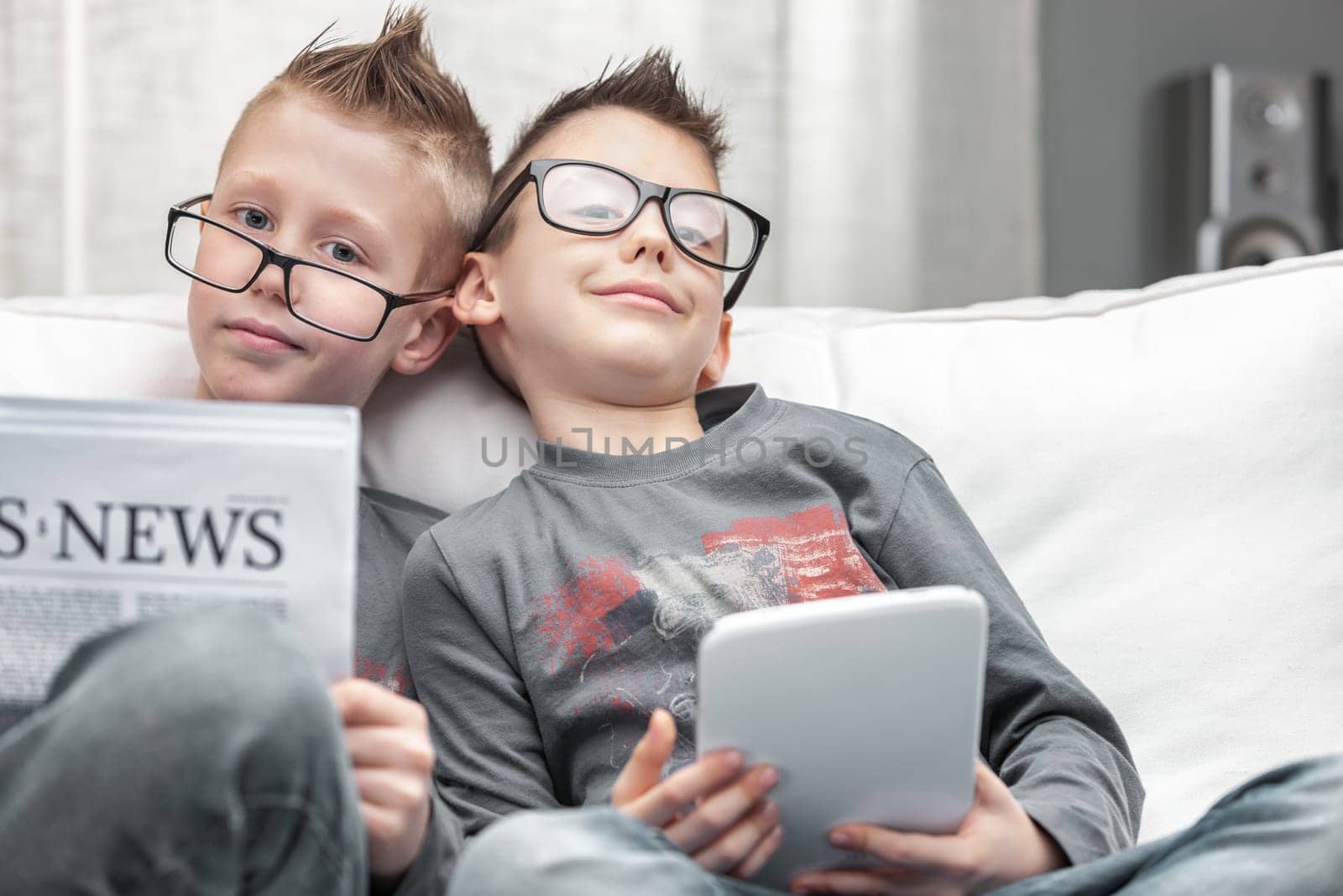 Two young boys wearing eyeglasses relaxing on a sofa together with tablet-pc and Business Times newspaper smiling happily at the camera