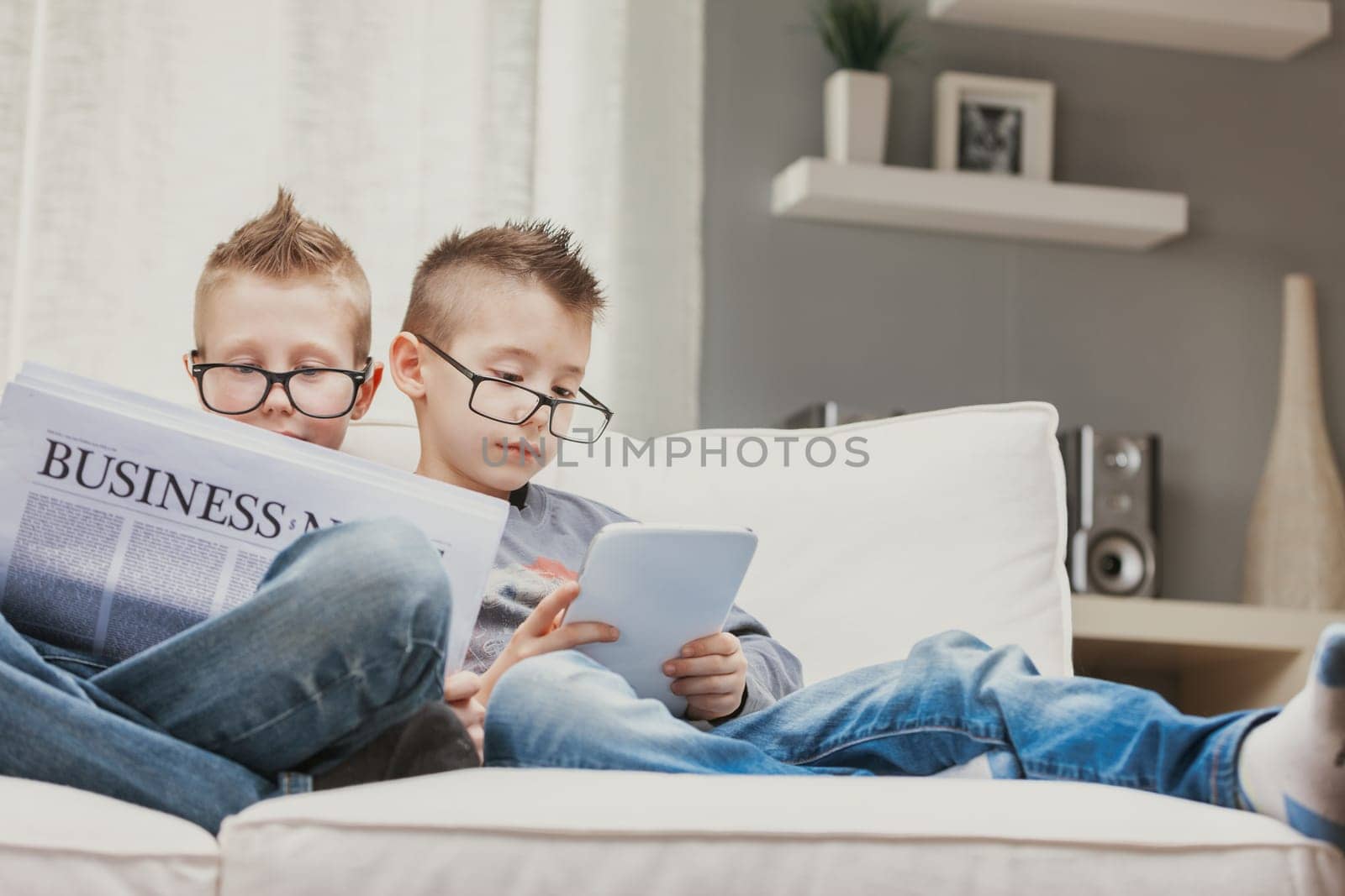 Two young boys wearing spectacles relaxing on a sofa reading the business news and an e-book on a tablet with engrossed expressions