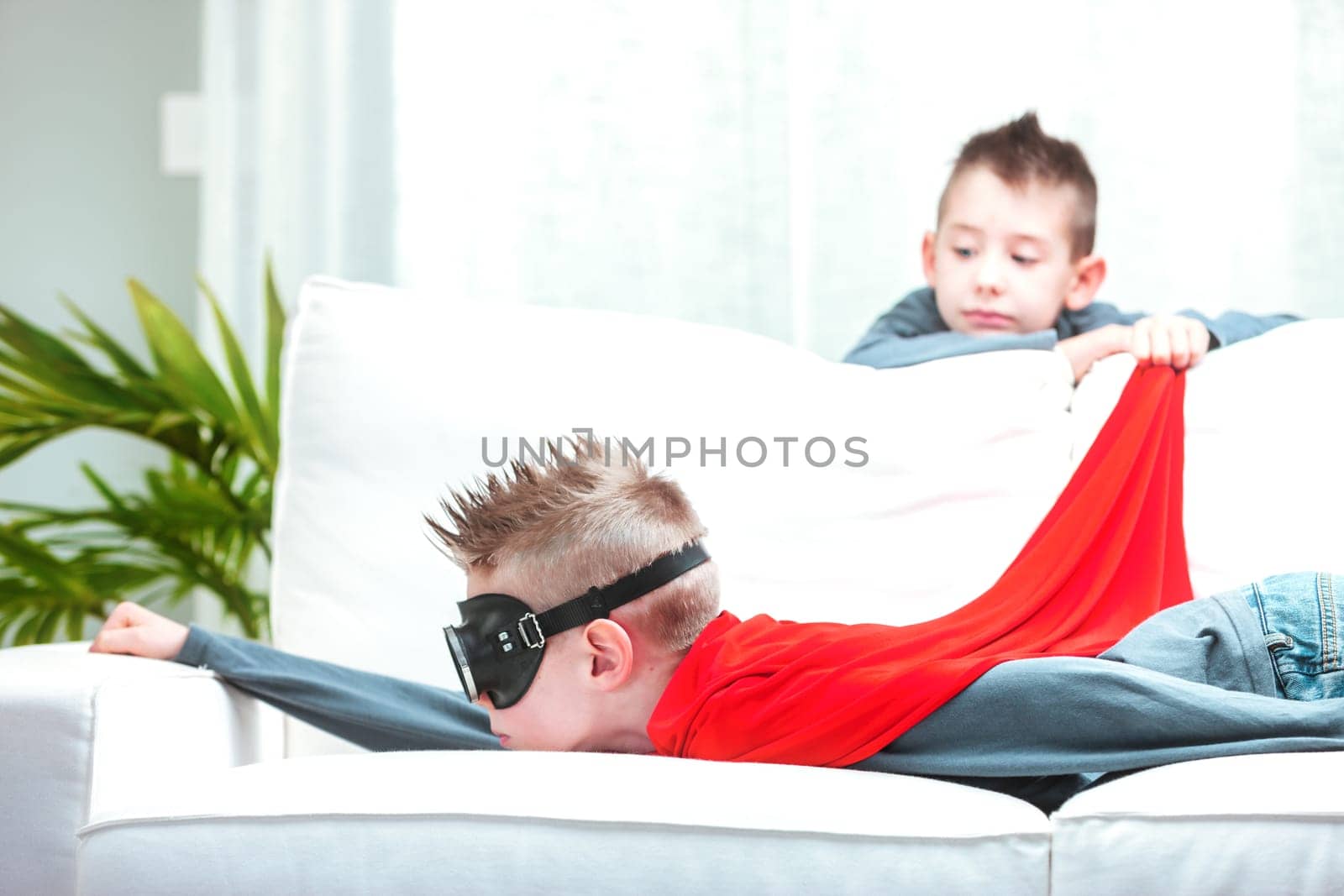 Young boy wth cute spiky hairstyle playing Super Hero as he mimics flying through the air with his red cape while lying on a sofa