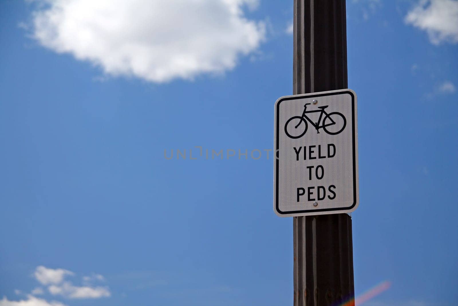 Yield to Peds sign hanging on a black pole with the sky and the clouds on the back, with bicycle icon