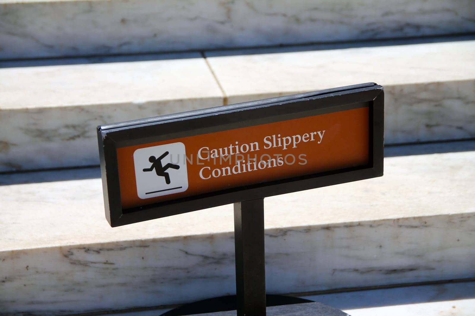 Caution, slippery conditions brown sign with icon representation on marble steps with a black holder on a sunny day