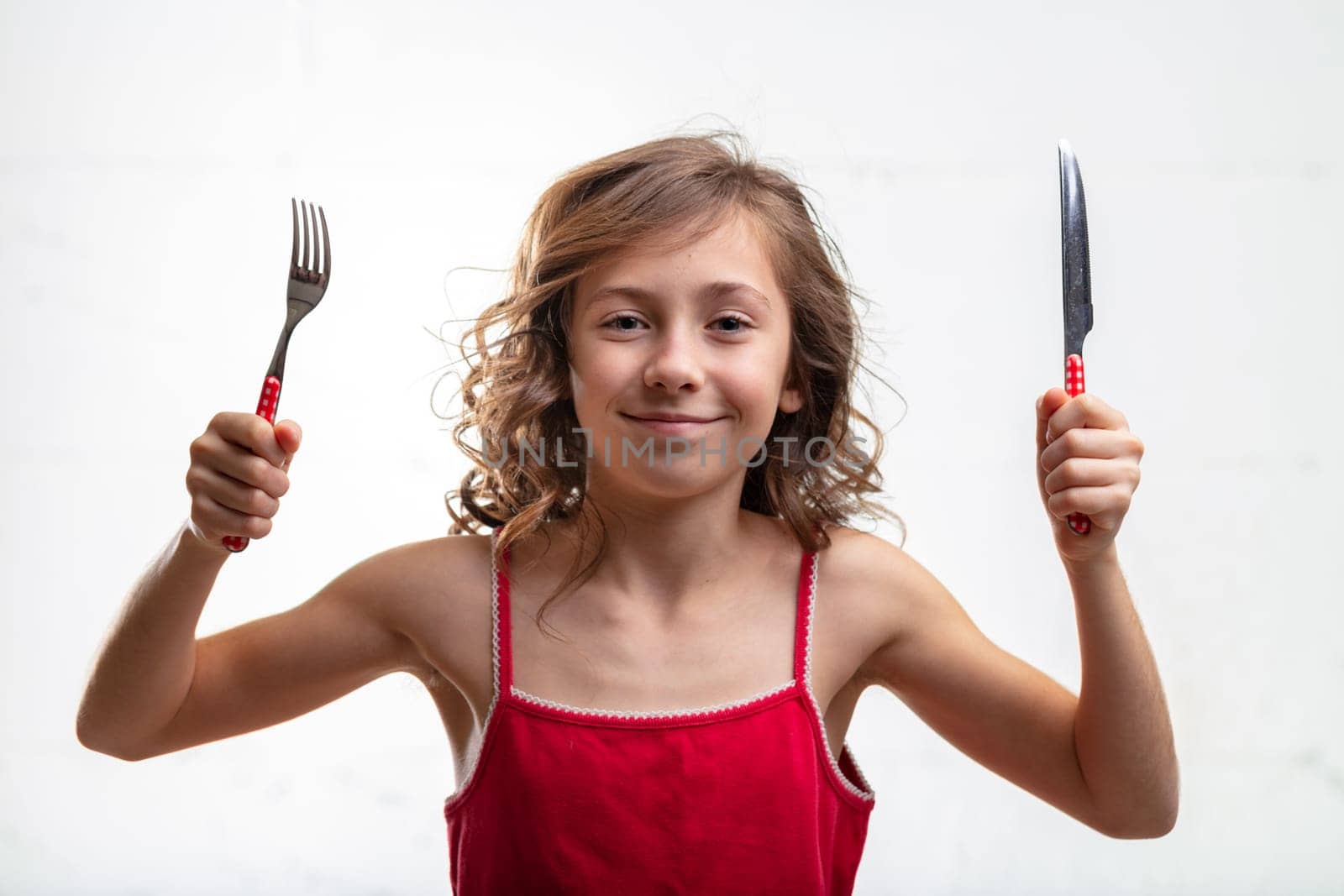 Playful young girl holding up a knife and fork in her hands with a mischievous smile isolated on white