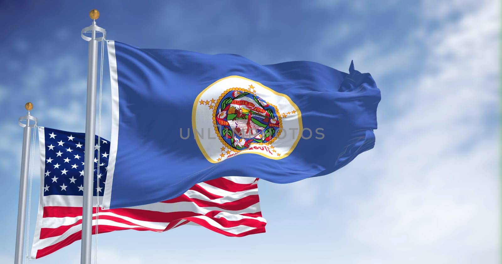 The Minnesota state flag waving along with the national flag of the United States of America. In the background there is a clear sky. Minnesota is a state in the upper Midwestern United States