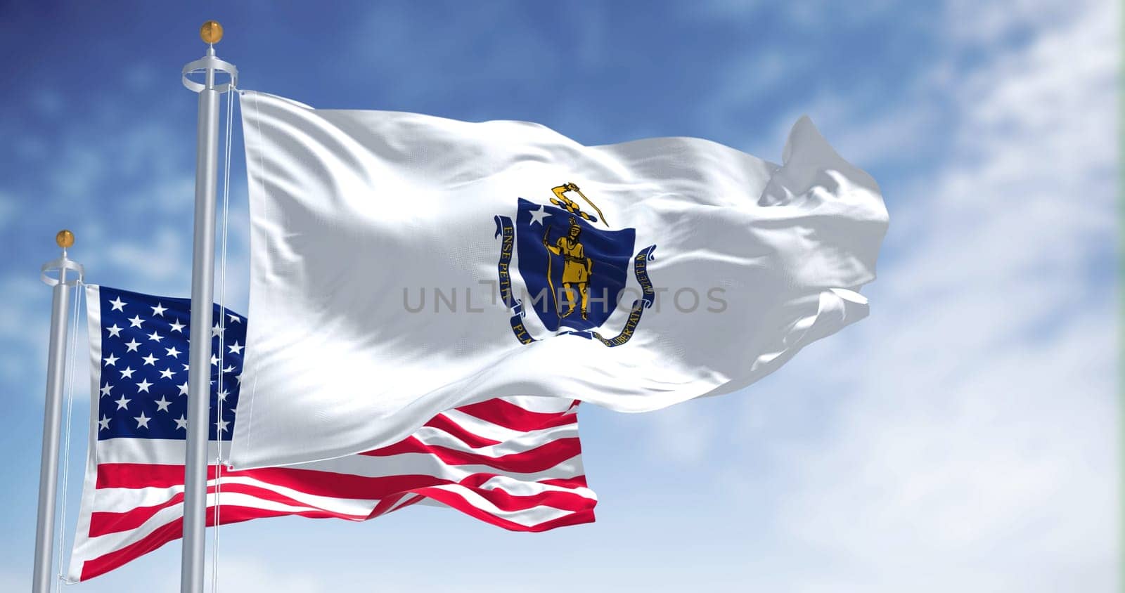 The Massachusetts state flag waving along with the national flag of the United States of America. In the background there is a clear sky. Massachusetts is a state in the New England region of the United States