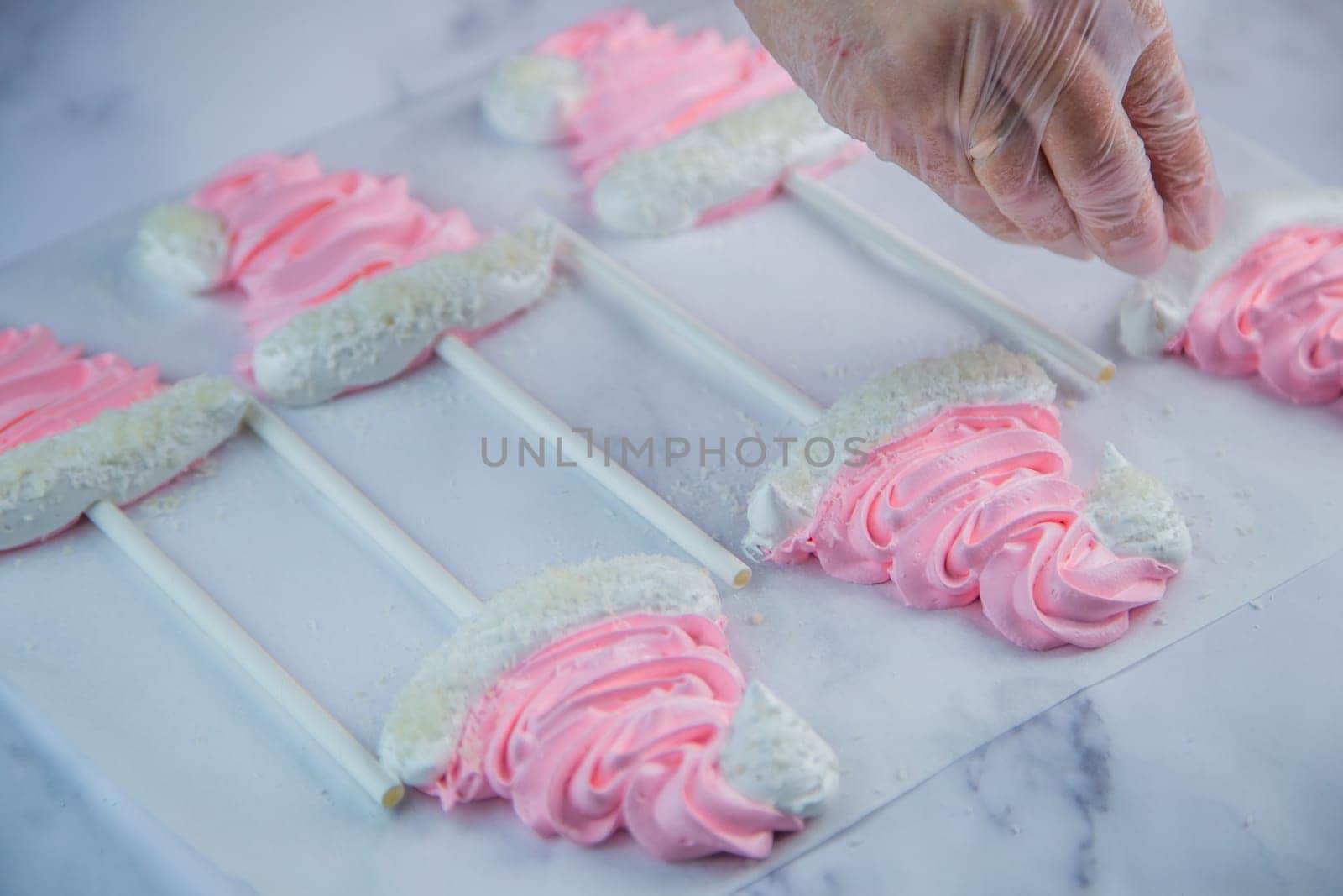 A pastry chef's hand in a transparent glove sprinkle coconut shavings on a pink meringue cap on a stick that lies on white parchment. High quality photo