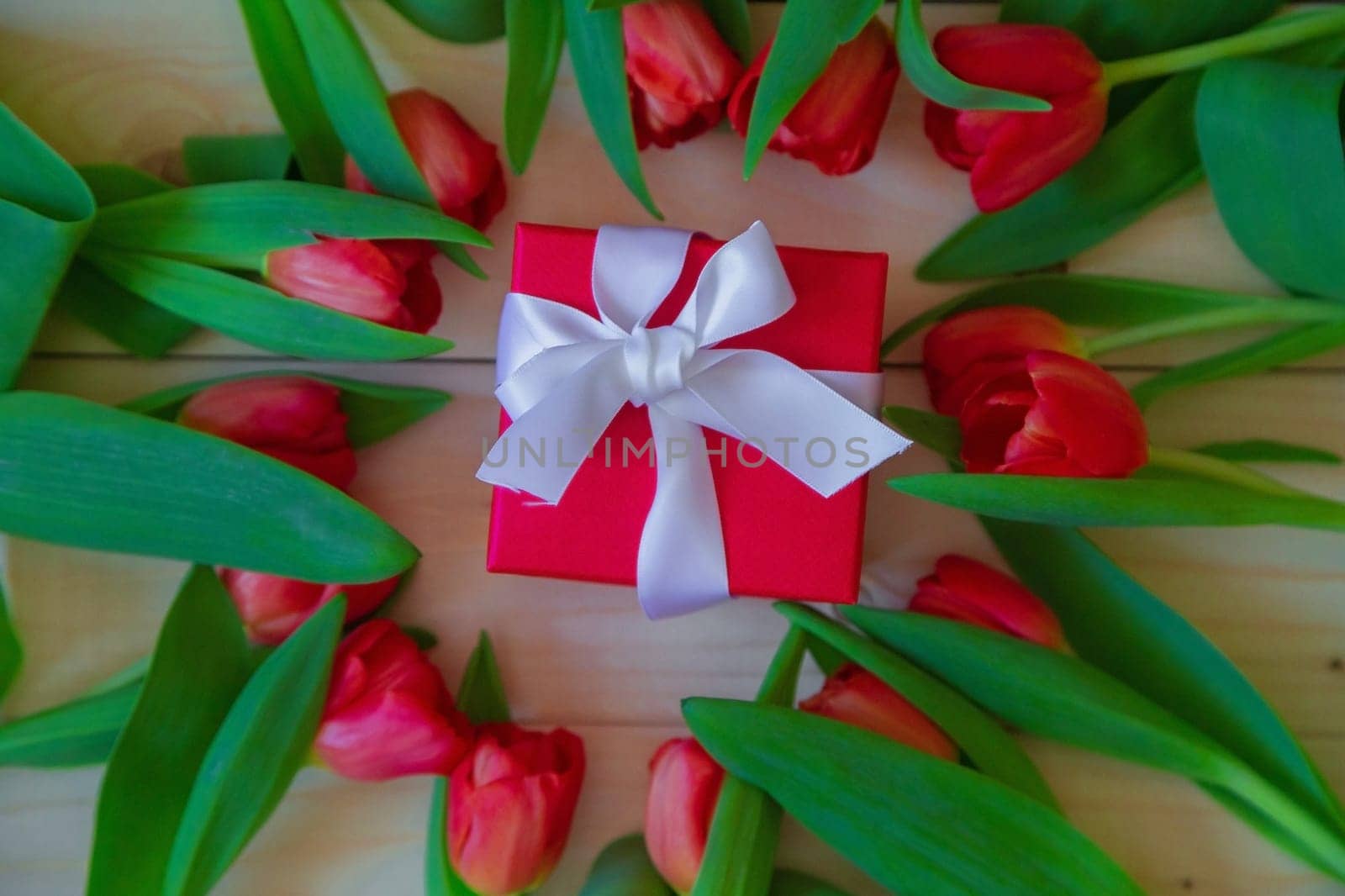 Pink tulips with green leaves lie in a circle on a wooden background, in the middle of Lesha's gift in a red package with a white bow. High quality photo