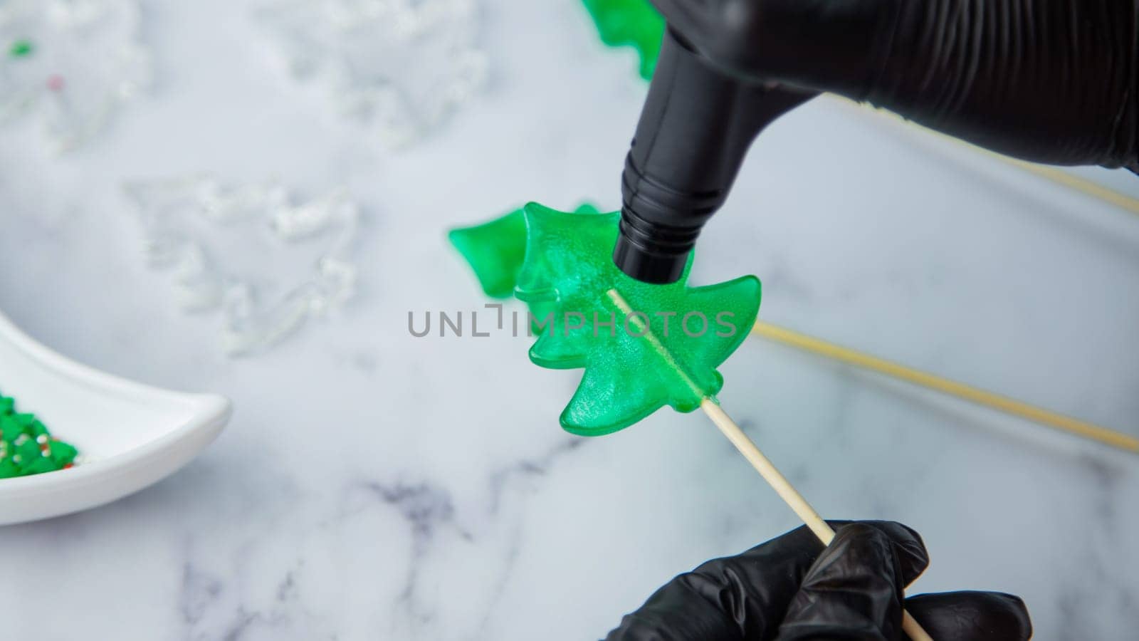 A green lollipop in the form of a Christmas tree is held in the hands of black gloves and burned on fire from a lighter, in the process of making a lollipop. High quality photo