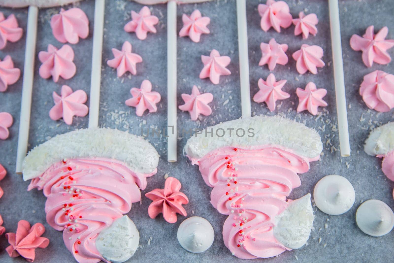 Meringues in the form of pink Christmas hats with a pompom and a white lapel on a stick lie on white parchment in two rows, pink stars and white meringues lie between them. High quality photo