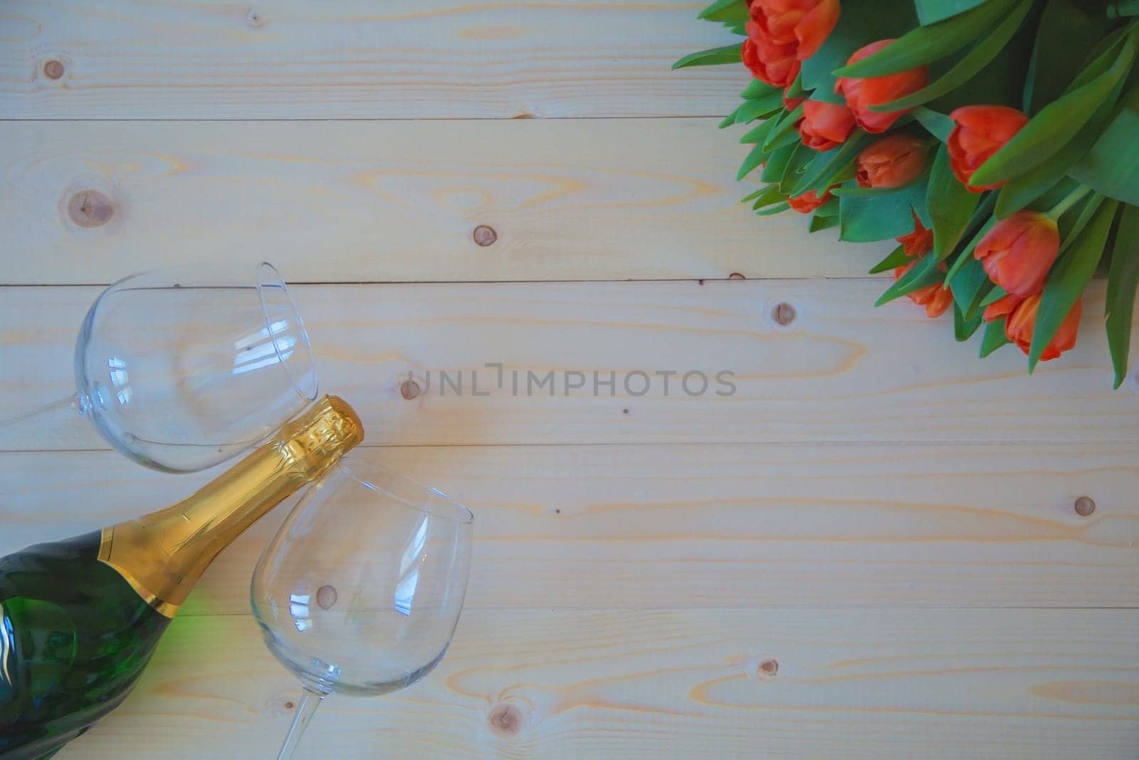 Pink tulips with green leaves lie in the upper corner on a wooden background, and opposite in the lower corner are wine glasses. High quality photo