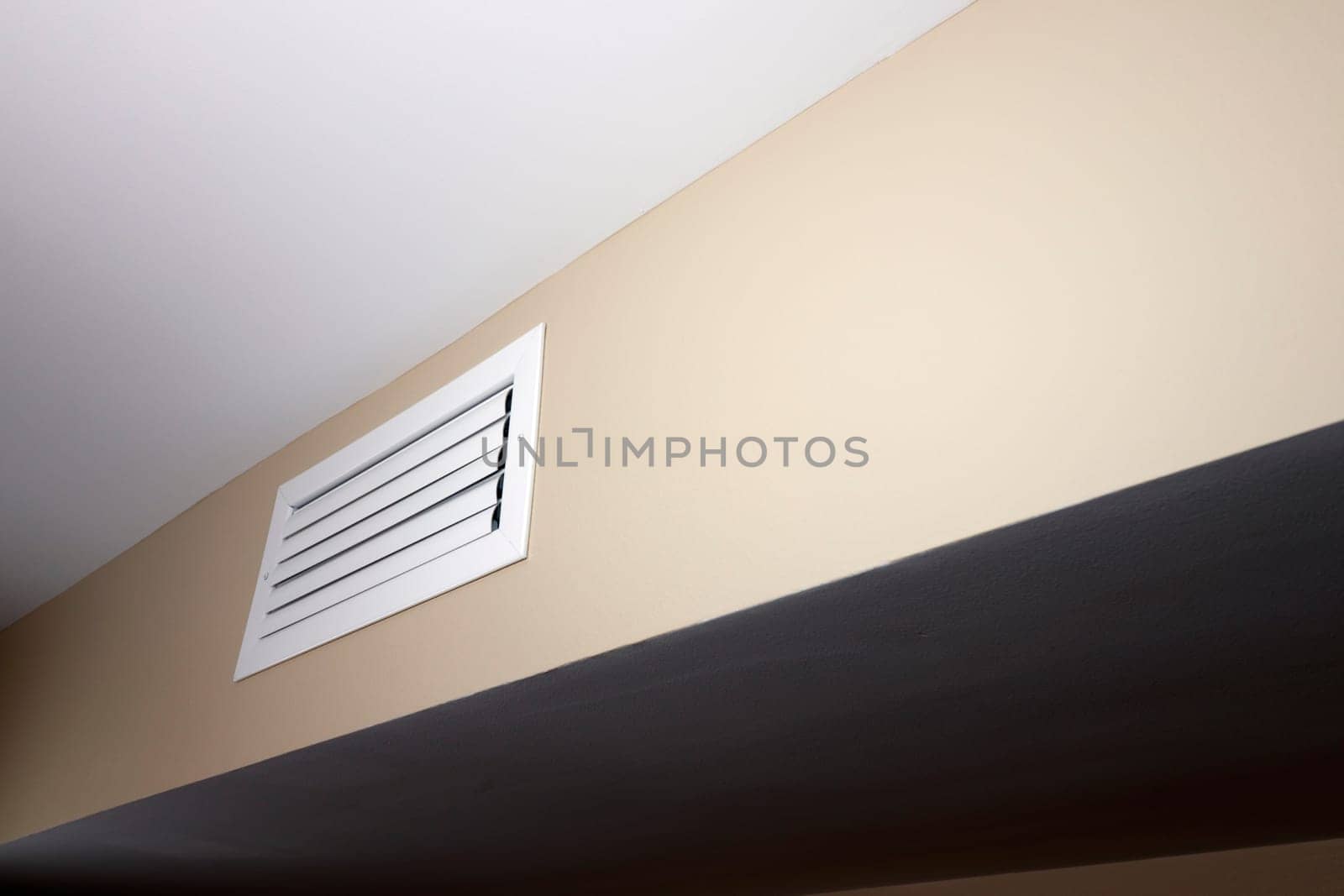 One closed air vent that is rectangle, white, small and situated in a beige wall near a white ceiling. A closed white rectangle air duct vent located in a home in a beige wall below a white ceiling.
