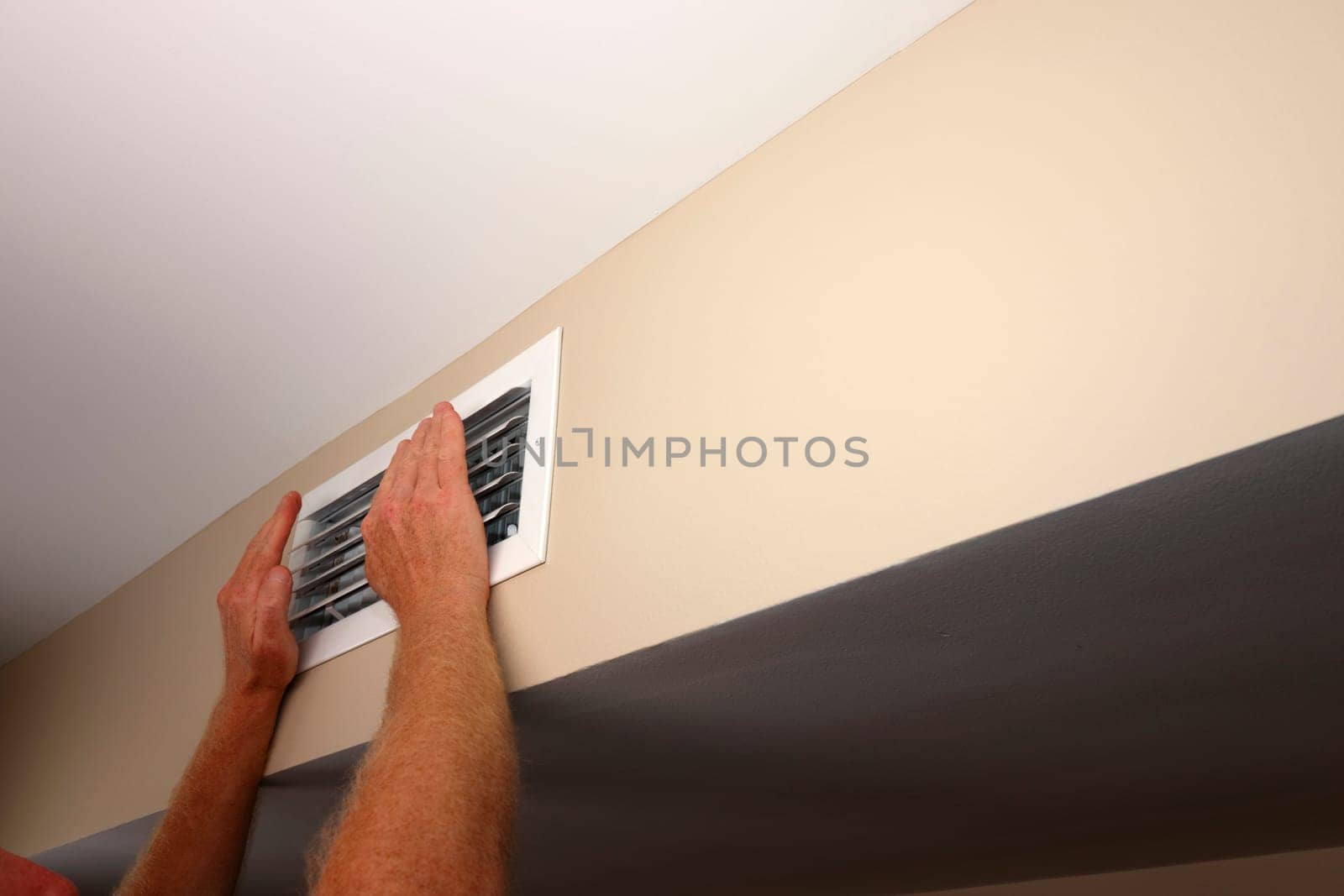 Feeling the air from a home air conditioning and heating HVAC vent on a wall are two hands in front of air duct grid. In a home near a ceiling is a rectangle white air vent with two hands in front of
