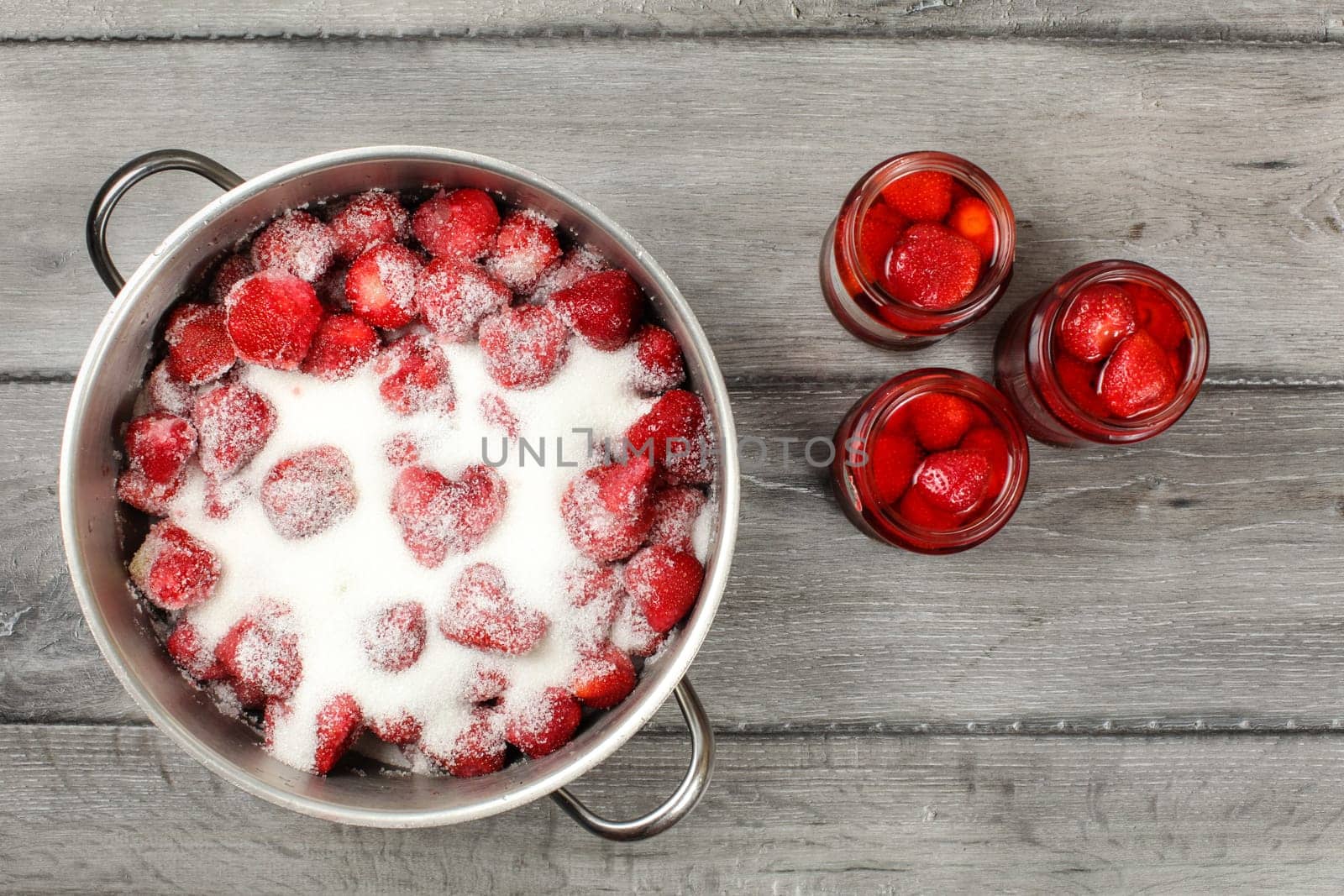 Tabletop view - large steel pot with strawberries covered in crystal sugar, three pickled strawberry glass bottles next to it. Homemade compote preparation.