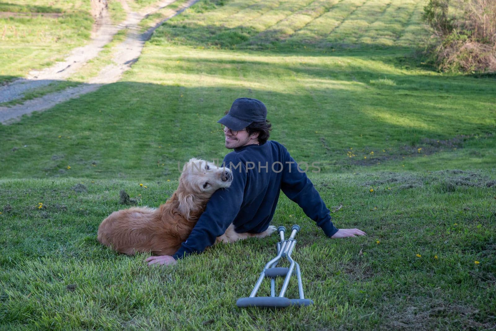 Dog cuddles with his owner on a rural grassy hill with a pair of crutches on the ground in the foreground. Dog looks back toward the camera in outdoor natural golden hour sunlight with nature background one person and copy space.