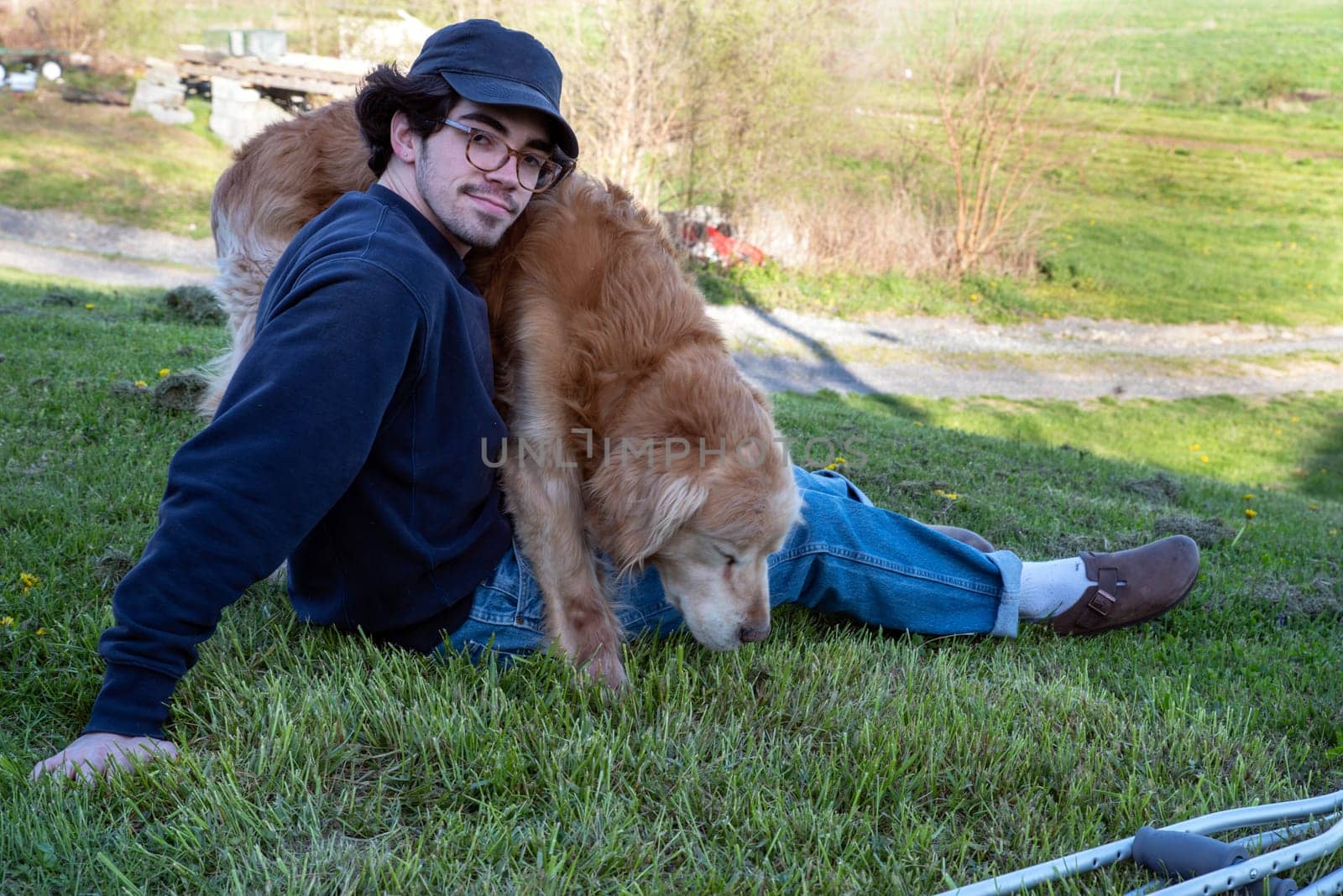 A handsome caucasian man sits alone on a grassy hill with a nature background and a loving golden retriever dog on his lap with a pair of crutches on the ground in the foreground. Selective focus, outdoors in natural light with copy space.