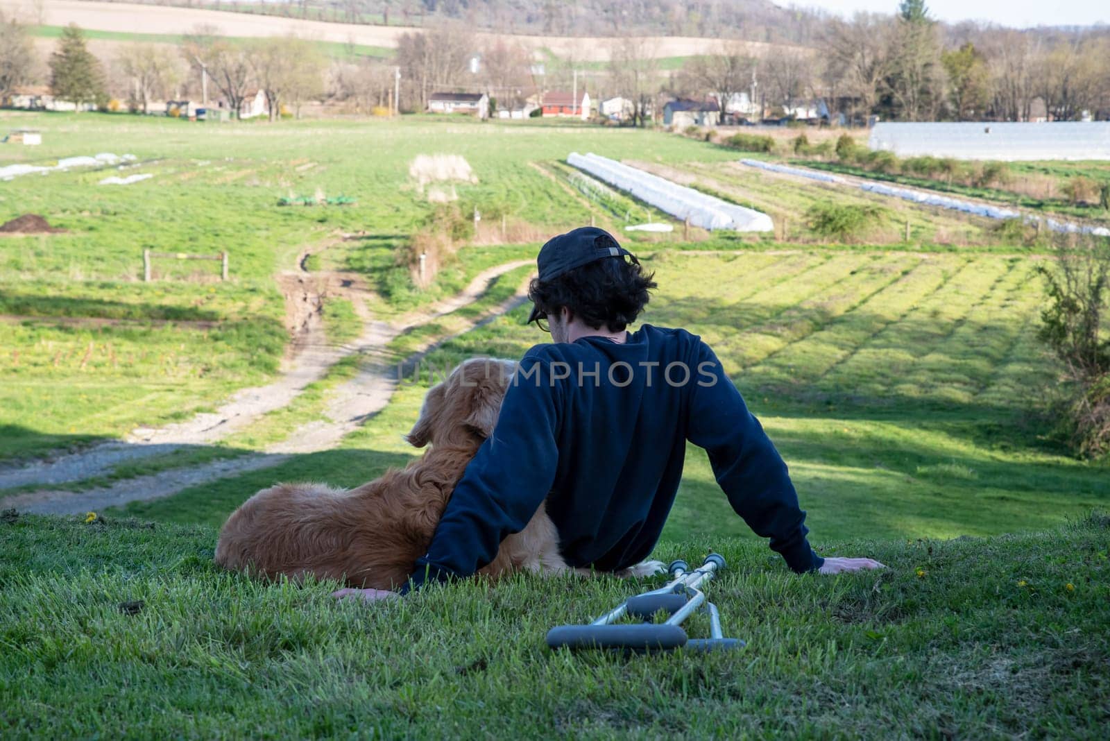 Crutches in the foreground of this idyllic rural farm background scene with a young man and his loving golden retriever dog cuddling with an arm around each other sitting on a grassy hill in natural golden hour sunlight one person and copy space.