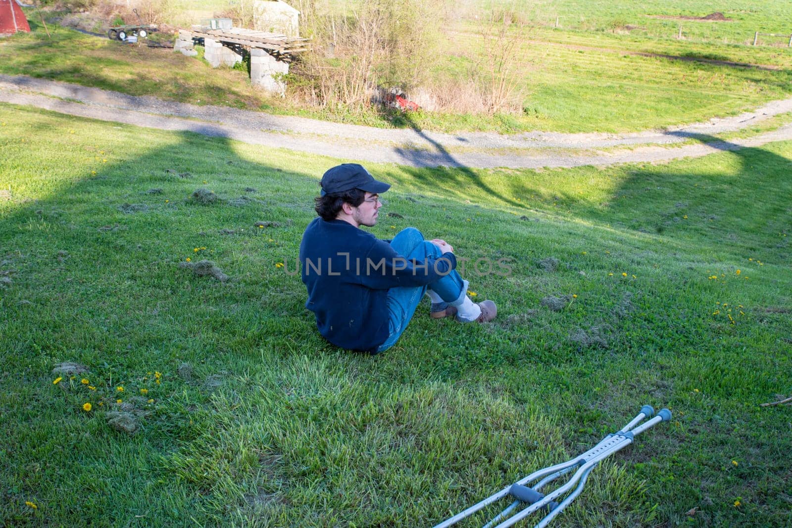 Young man sits alone on a grassy hill in a rural farm landscape with dirt road in background and a pair of crutches in the foreground laying on the ground. Serene image with shadow outline of a house in golden hour sunlight with copy space.