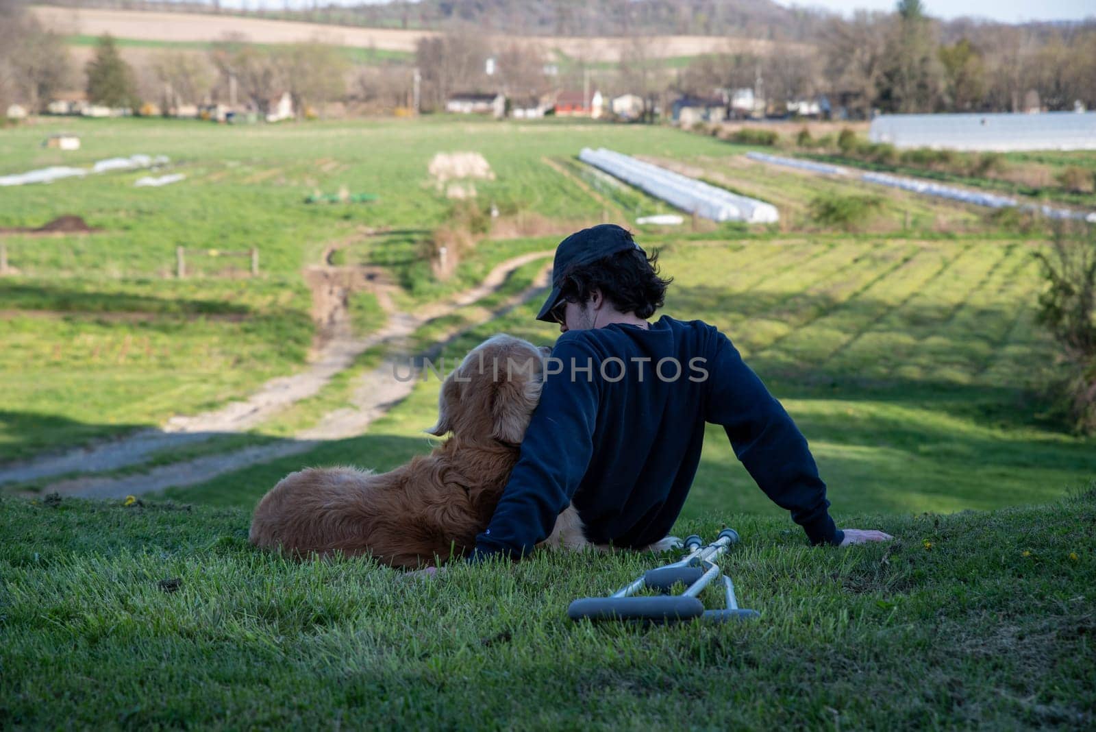 Crutches in the foreground of this idyllic rural farm background scene with a young man and his loving golden retriever dog cuddling and looking at each other sitting on a grassy hill in natural golden hour sunlight one person and copy space.