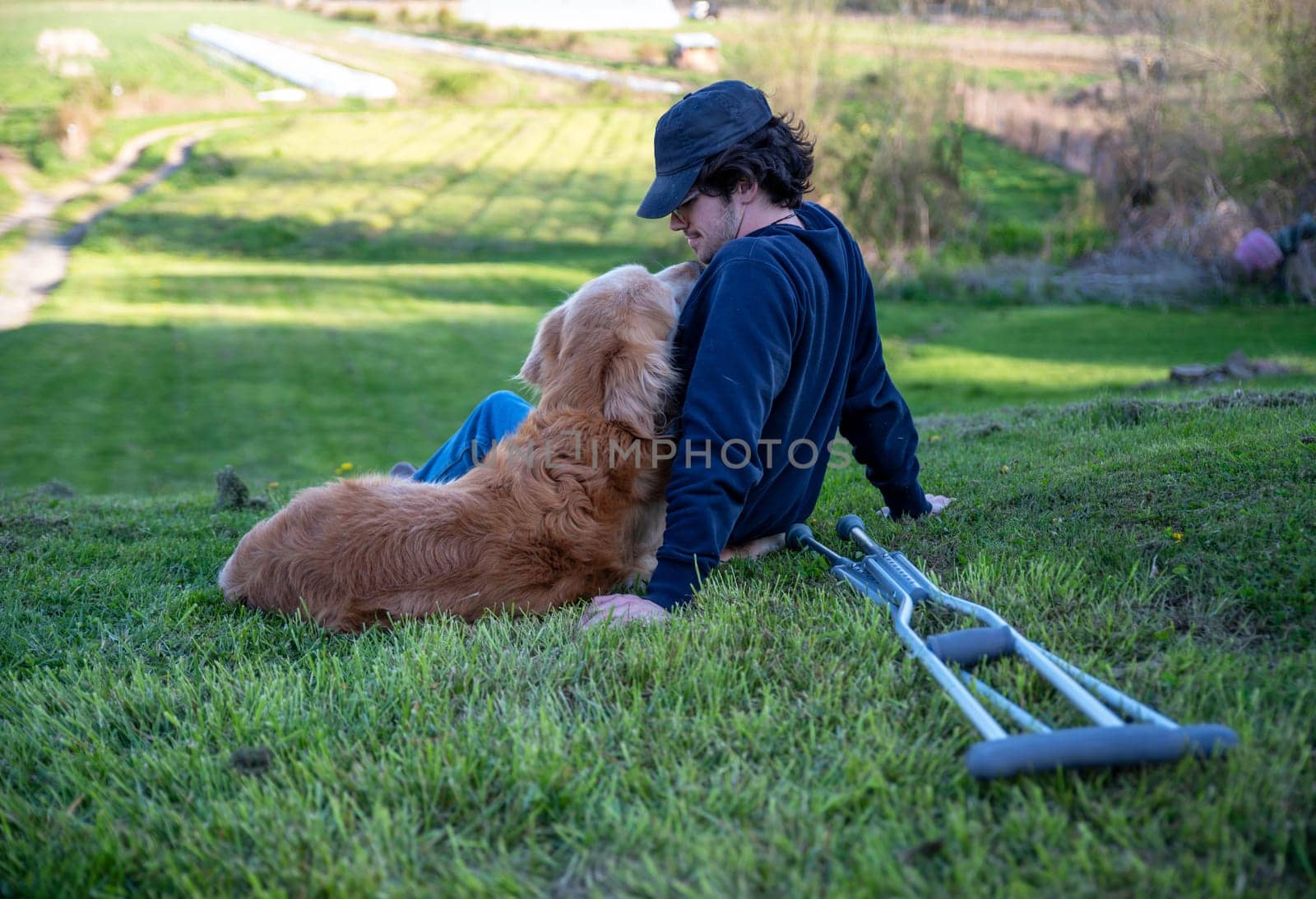 Crutches in the foreground of this idyllic rural farm background scene with a young man and his loving golden retriever dog cuddling with an arm around each other sitting on a grassy hill in natural golden hour sunlight one person and copy space.