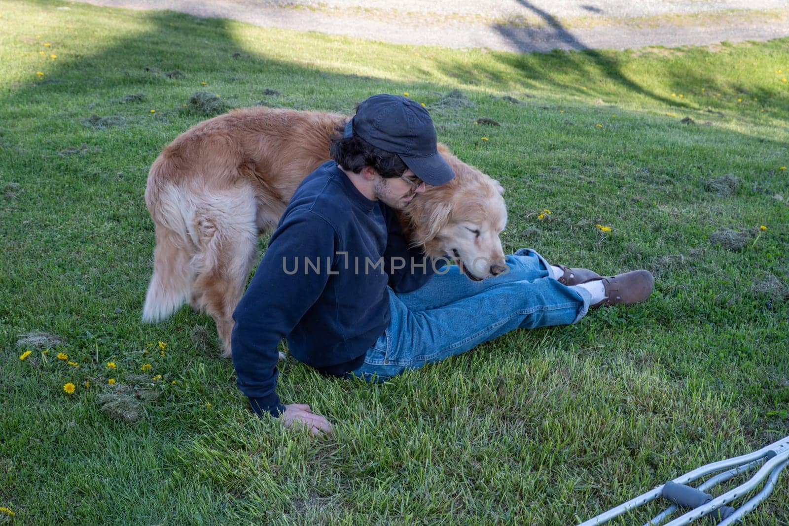 Handsome young man sits on a grassy hill with crutches laying in the foreground. A large loving Golden Retriever dog cuddles with him. Animal companionship with a green nature background shot in natural light with copy space.