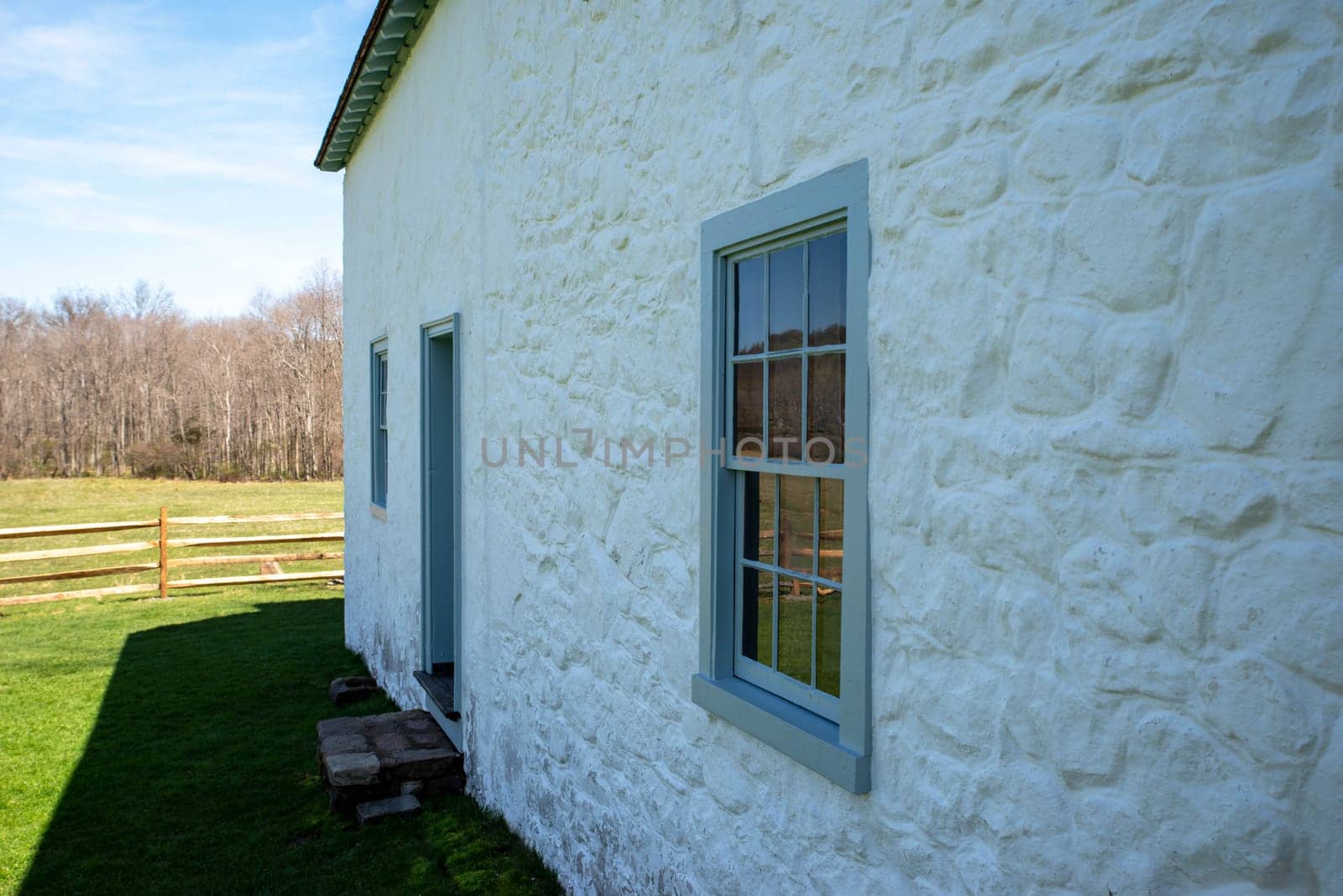 Beautiful sude view of a whitewashed stone cottage with blue window rim and antique window panes with rural scene reflection. Stone texture and green pasture and woodland background in late afternoon natural light with copy space and no people.