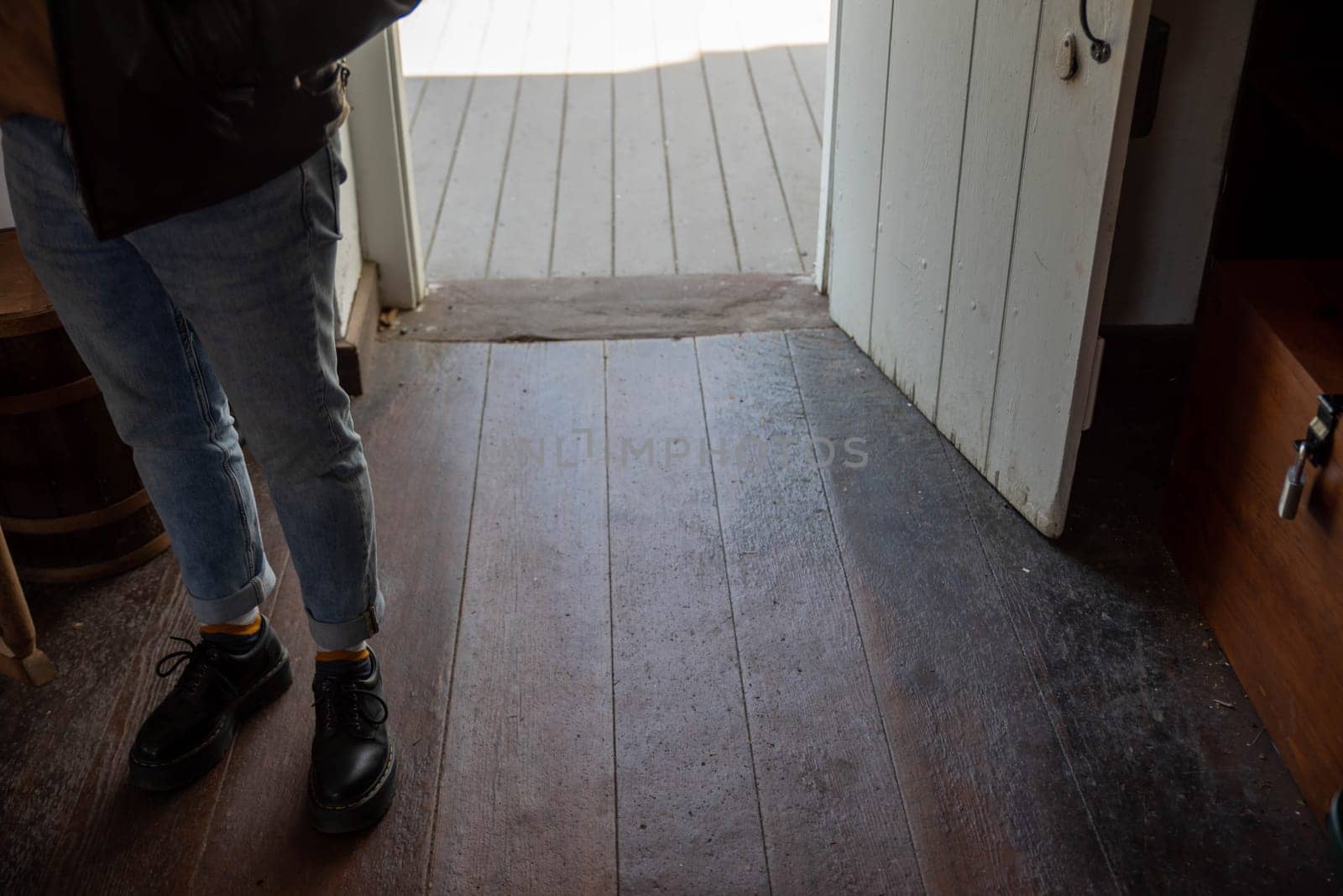 One person's feet stand inside the open door of an old colonial American home with old, hardwood floors and rustic antique door and stone threshold. Selective focus on foreground with copy space and wood grain texture.