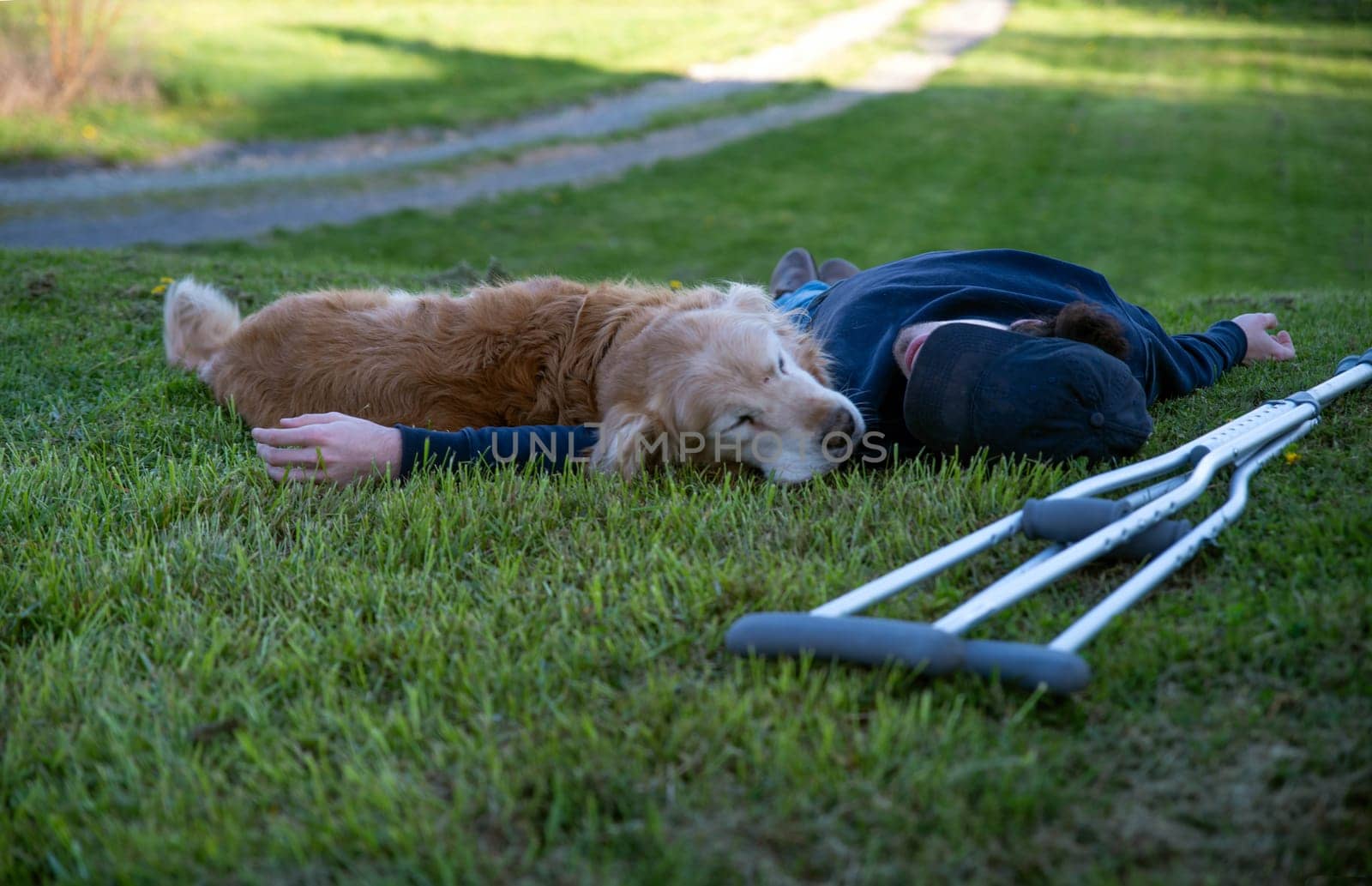 Crutches in the foreground of this idyllic rural nature background scene with a young man and his loving golden retriever dog laying on a grassy hill in natural golden hour sunlight one person and copy space.
