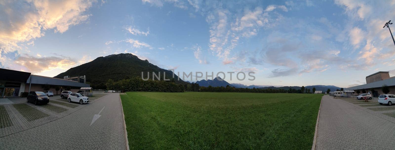 panoramic view of a modern mountain town in northeastern Italy, with the blue sky mottled with clouds and a beautiful green lawn