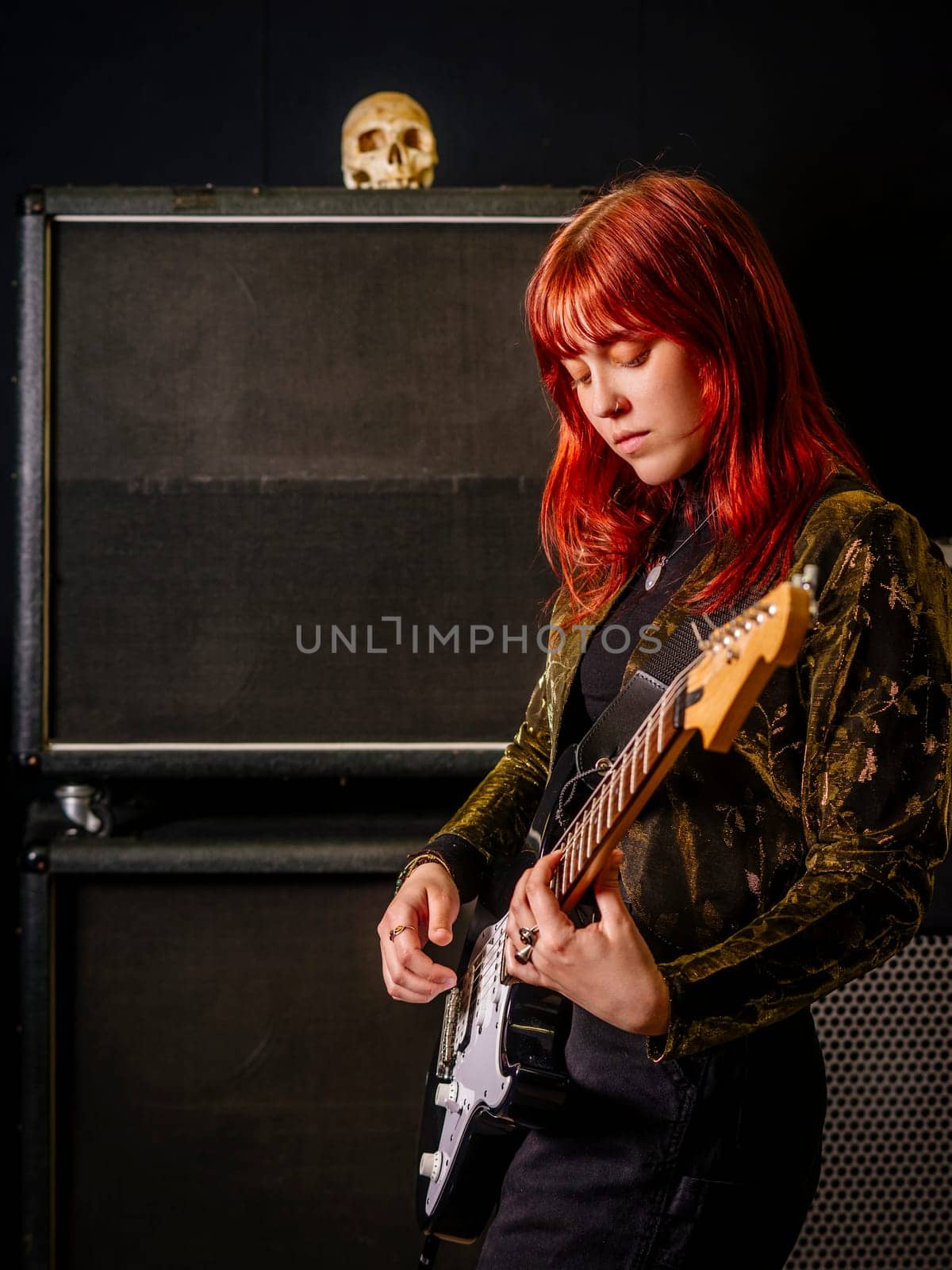Young redhead woman playing electric guitar in front of large speaker cabinets.