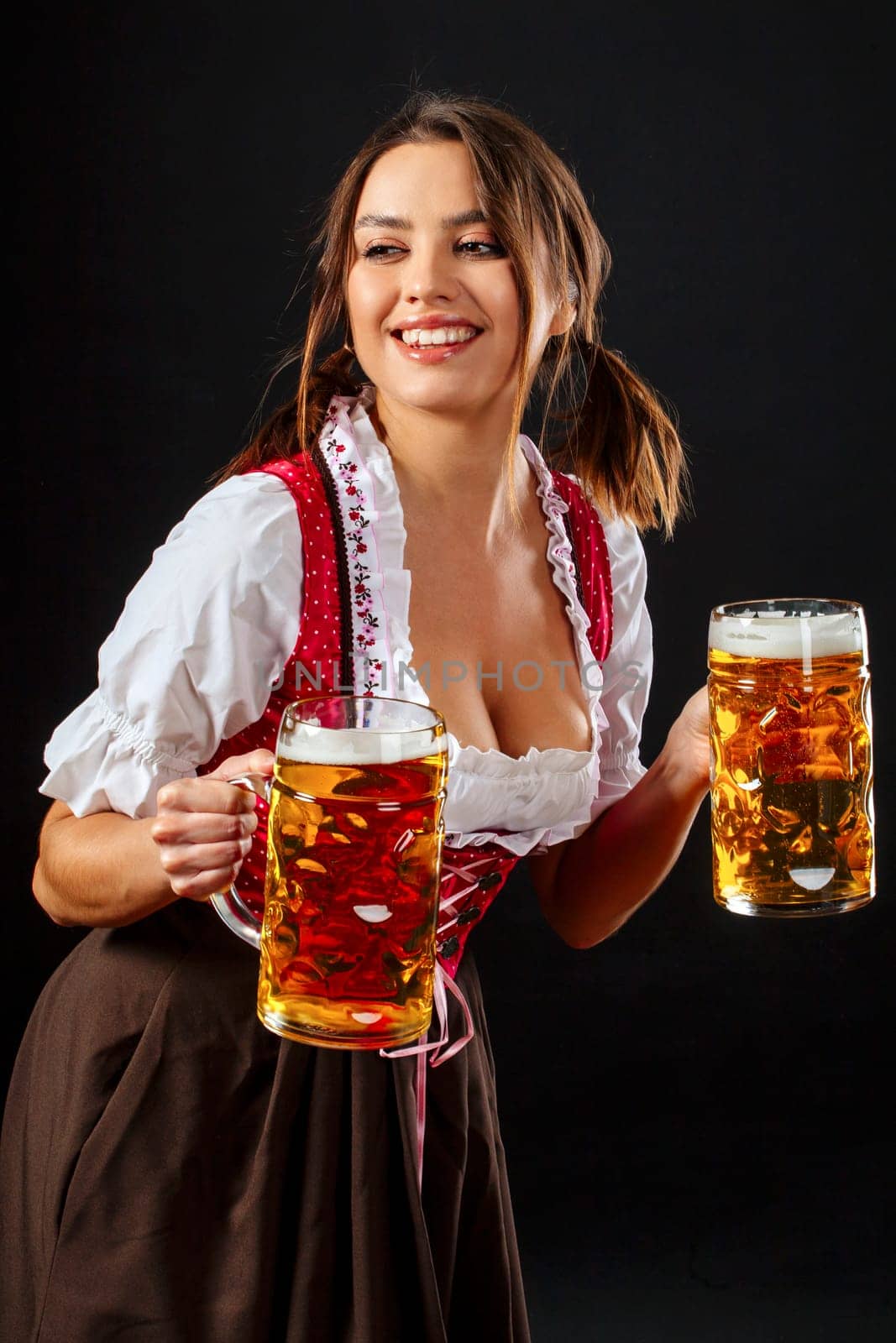 Beautiful waitress wearing traditional dirndl and holding two mass beer steins over black background.