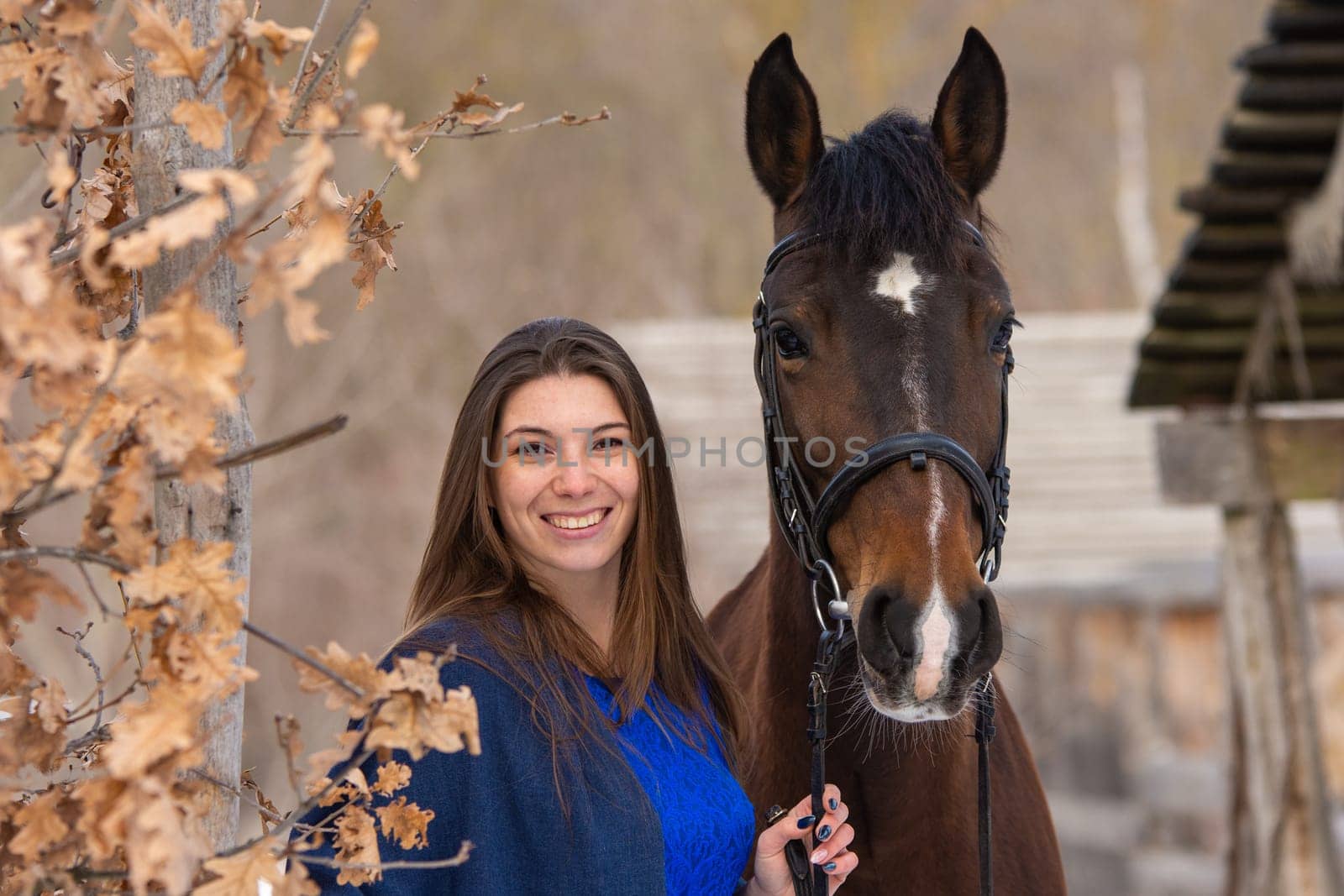 Close-up portrait of a horse and a beautiful girl of Slavic appearance, against the backdrop of a winter forest and an old farm
