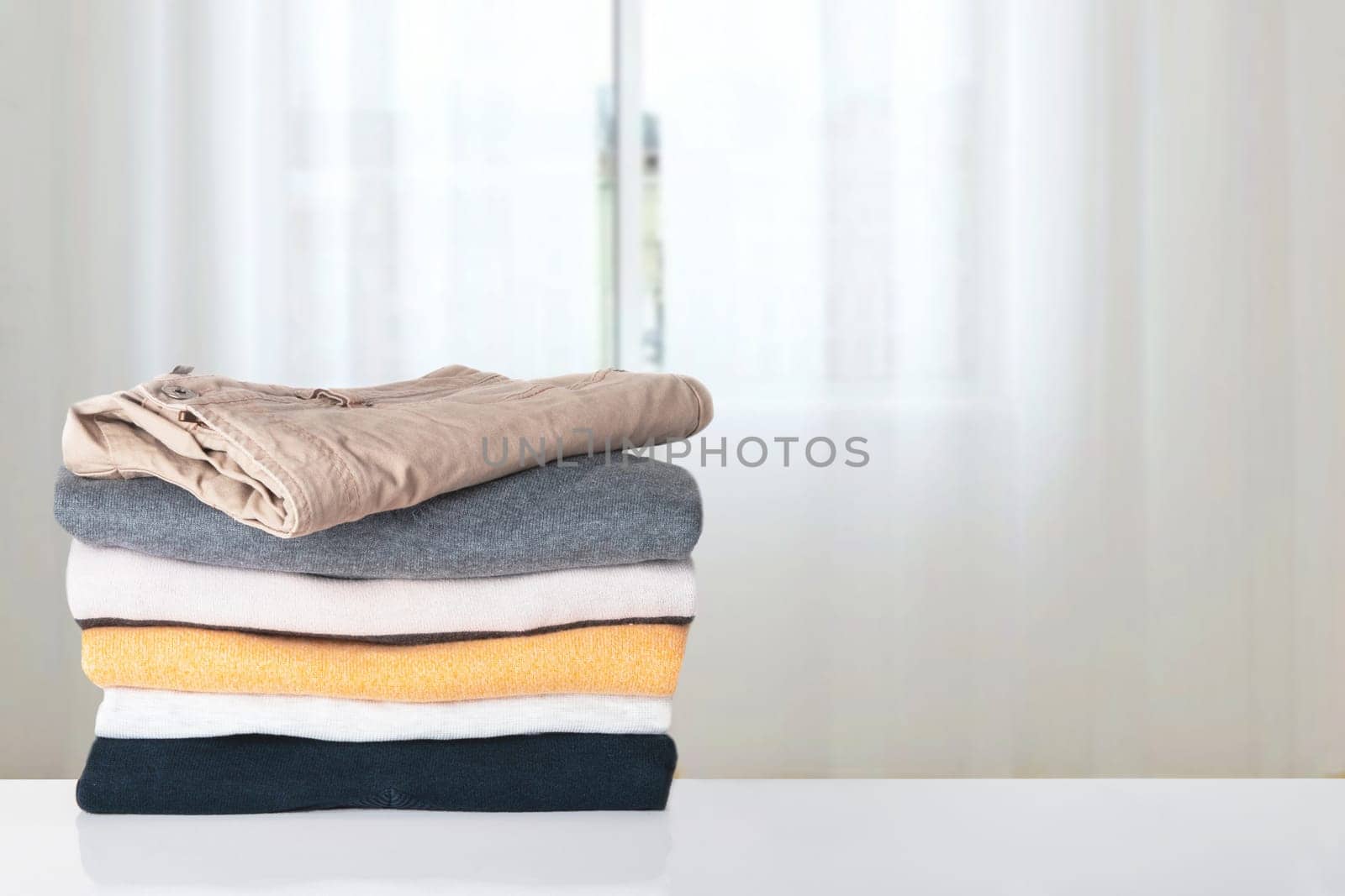 Stack of spring clothes. Pile of colorful knitted cozy warm cashmere sweaters or pullover on table against abstract bright curtain background. Space for display product montage. Beautiful backdrop.