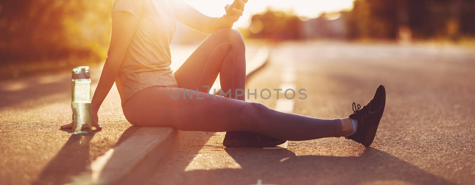 Woman sitting on the border of the roadside with smartphone after a run. Panoramic view. Concept of the healthy lifestyle.