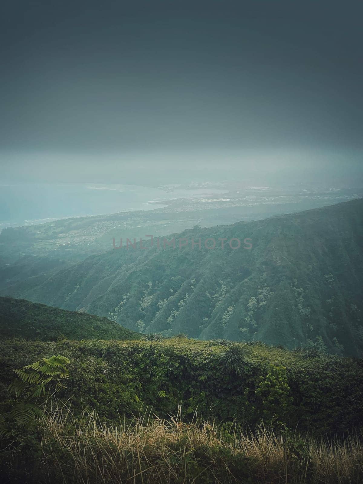 Scenery view from the top of the mountains to the ocean coastline in Hawaii, Oahu island. Moody hiking landscape with foggy, green hills on the horizon and stormy clouds over the bay