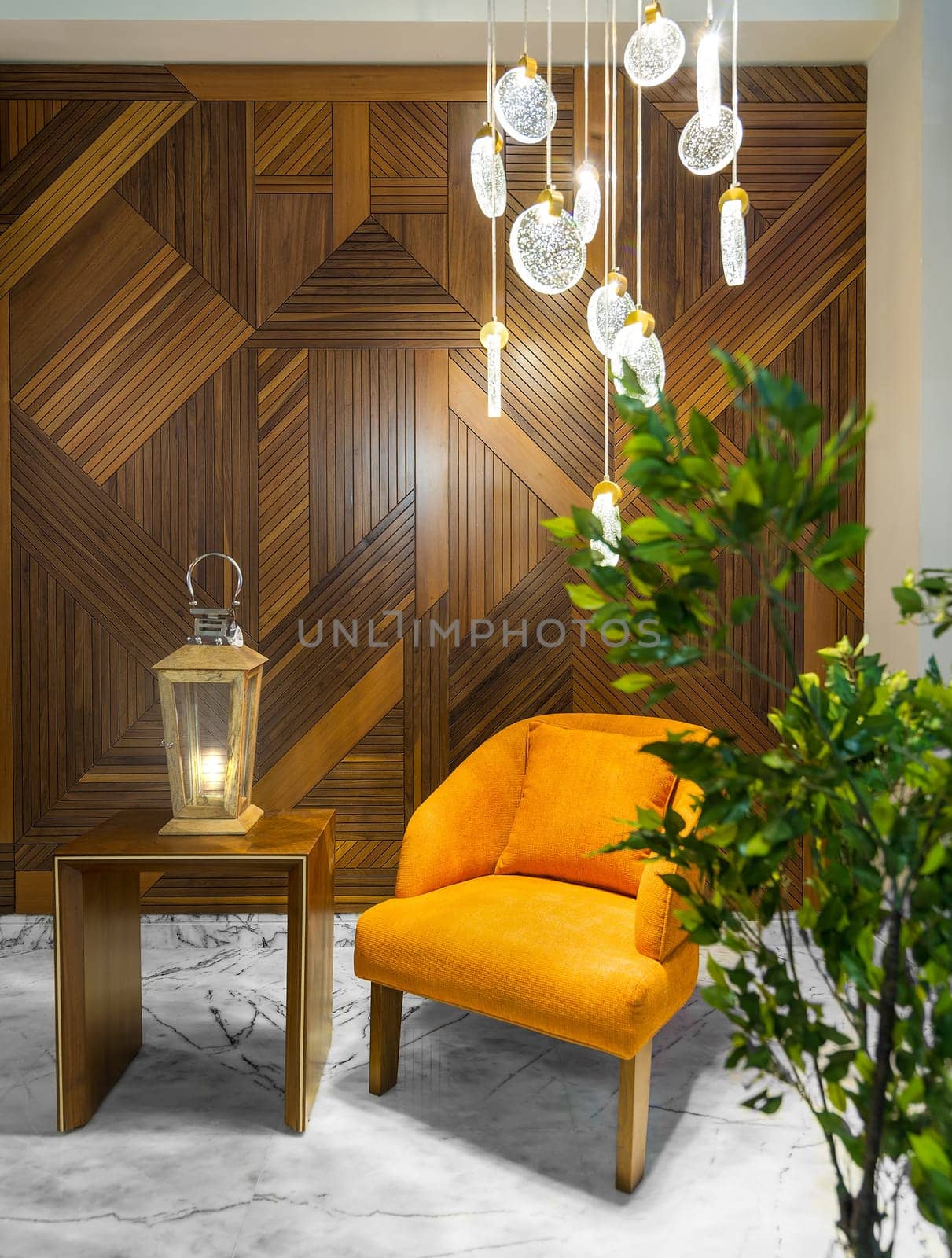 Orange armchair, illuminated wooden lantern on small wooden table, and contemporary tall glass chandelier, in a hall with decorated wood cladding wall, and white marble floor