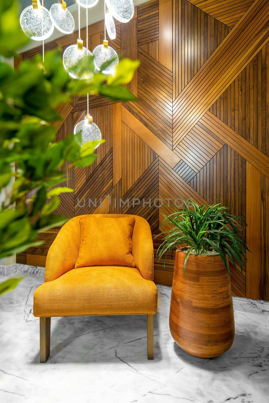 Modern orange armchair, tall wooden planter, and contemporary glass chandelier, in a hall with decorated wood cladding wall, and white marble floor, framed by green leaves