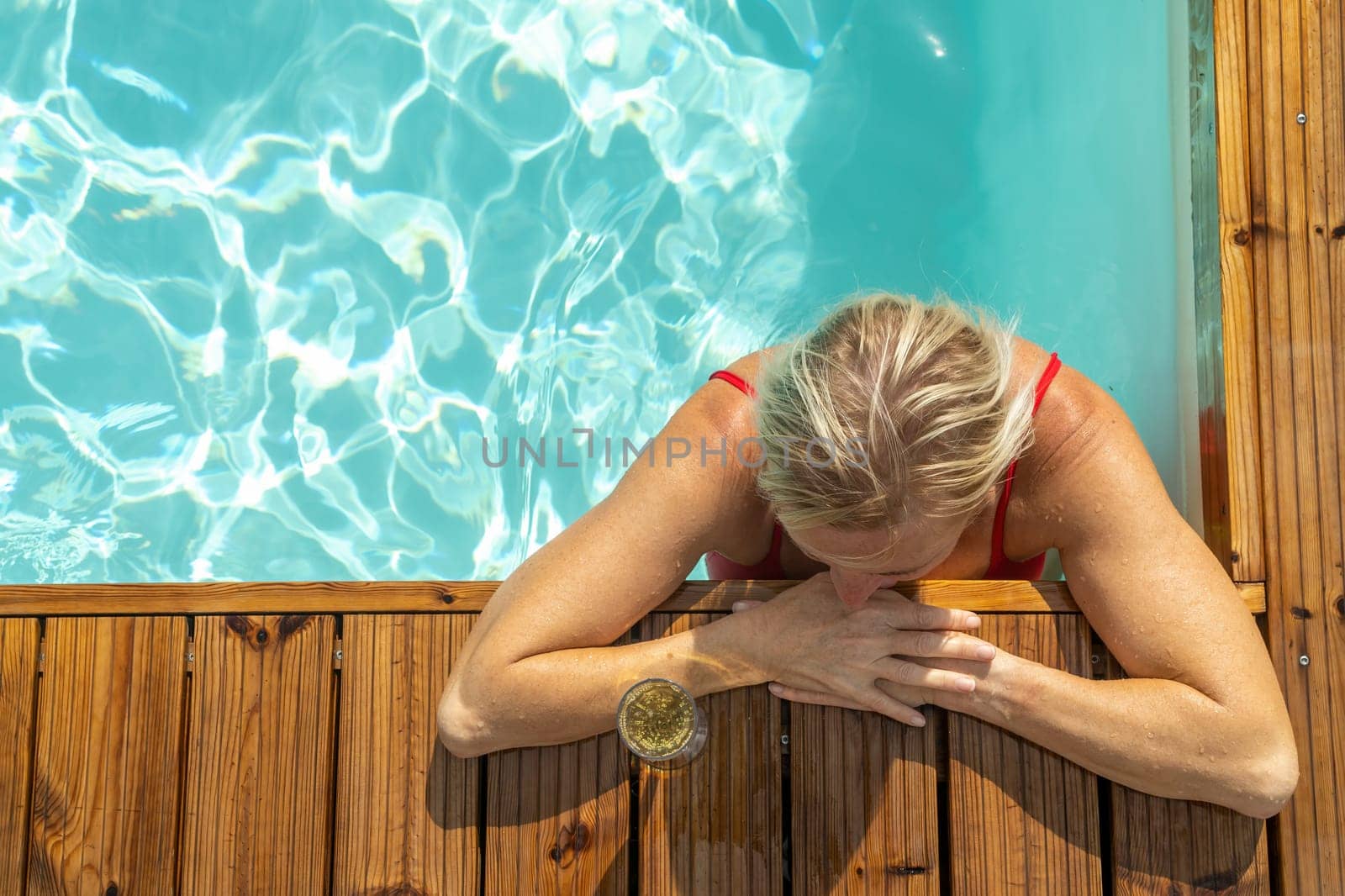 Top view of wet woman standing in swimming pool and leaning on wooden edge.