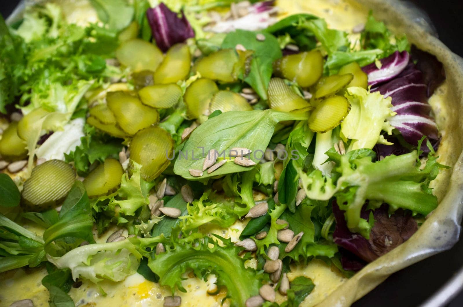 Hot salad over .scrambled egg on pan. Plate with several lettuces, gherkins and sunflowers seeds