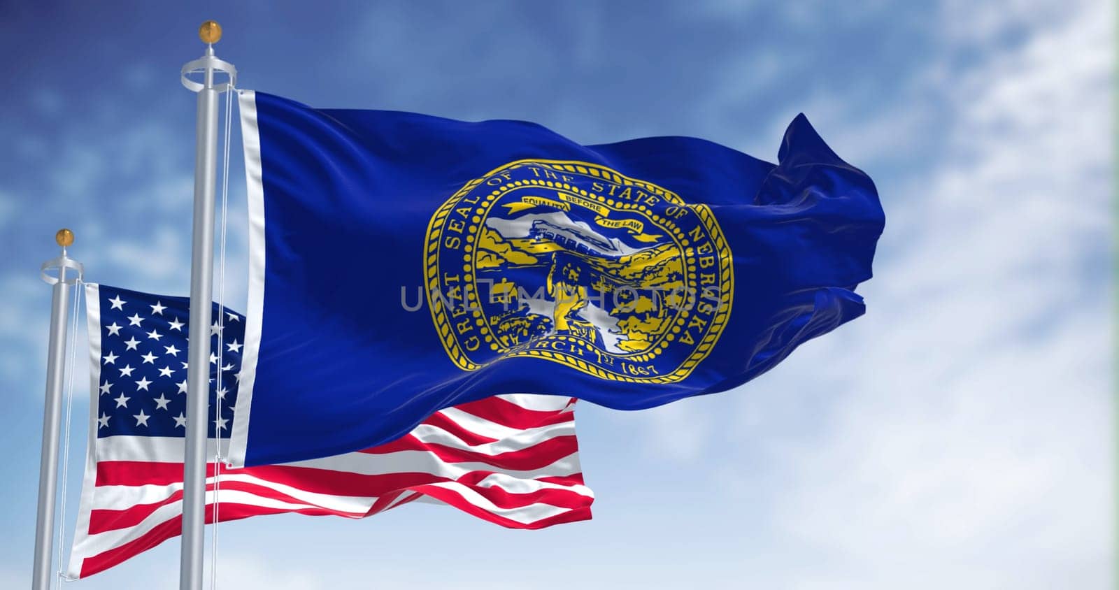 The Nebraska state flag waving along with the national flag of the United States of America. In the background there is a clear sky. Nebraska is a state in the Midwestern United States