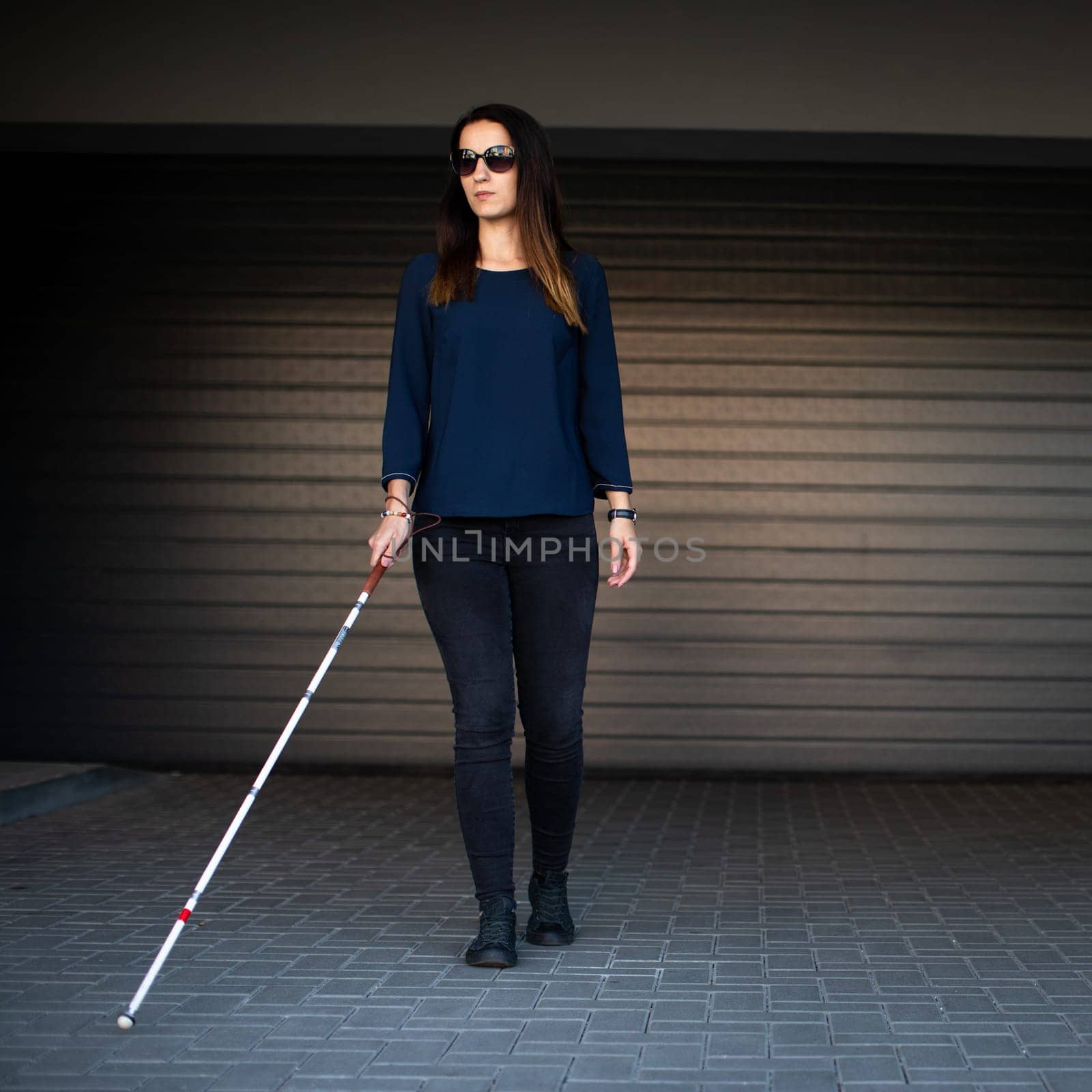 Young woman with impaired vision walking on city streets, using her white cane to navigate the urban space better and to get to her destination safely