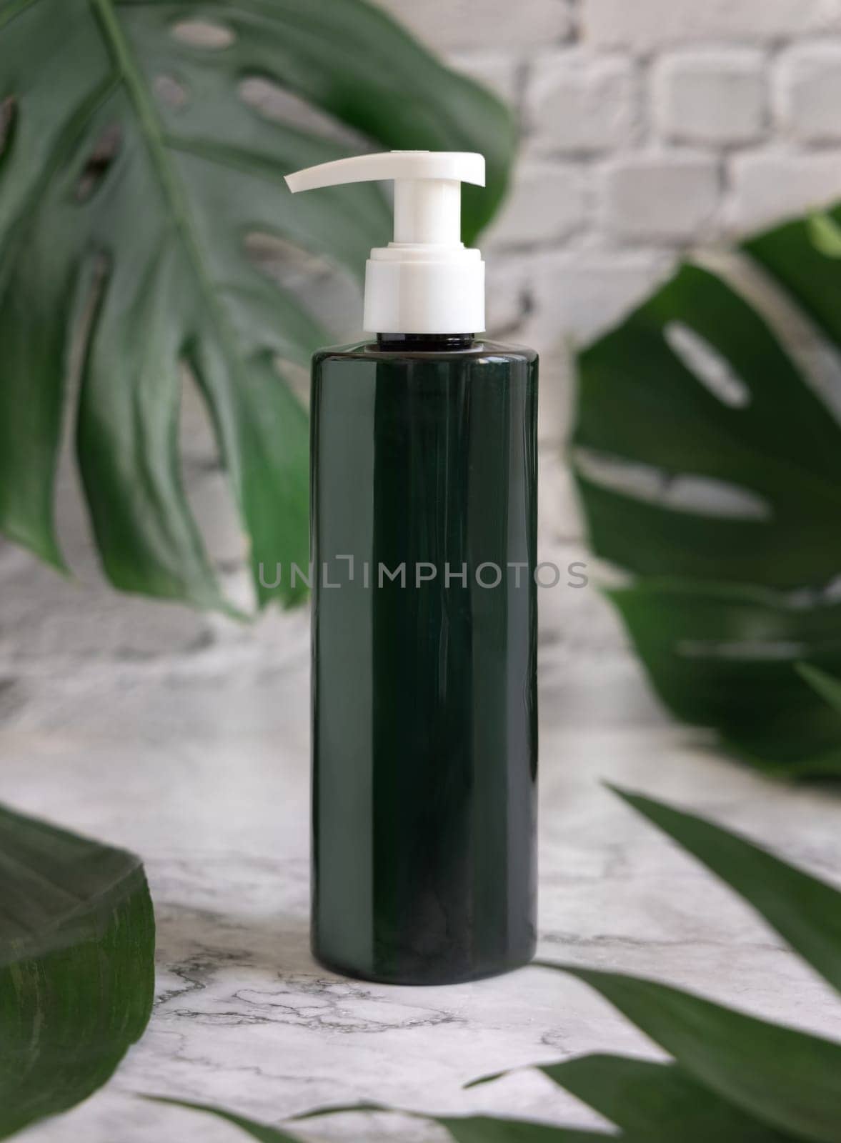 Dark green one pump dispenser and tropical monstera leaves on marble table close up. Brand packaging mockup. Skincare beauty product. Natural herbal cosmetics concept..