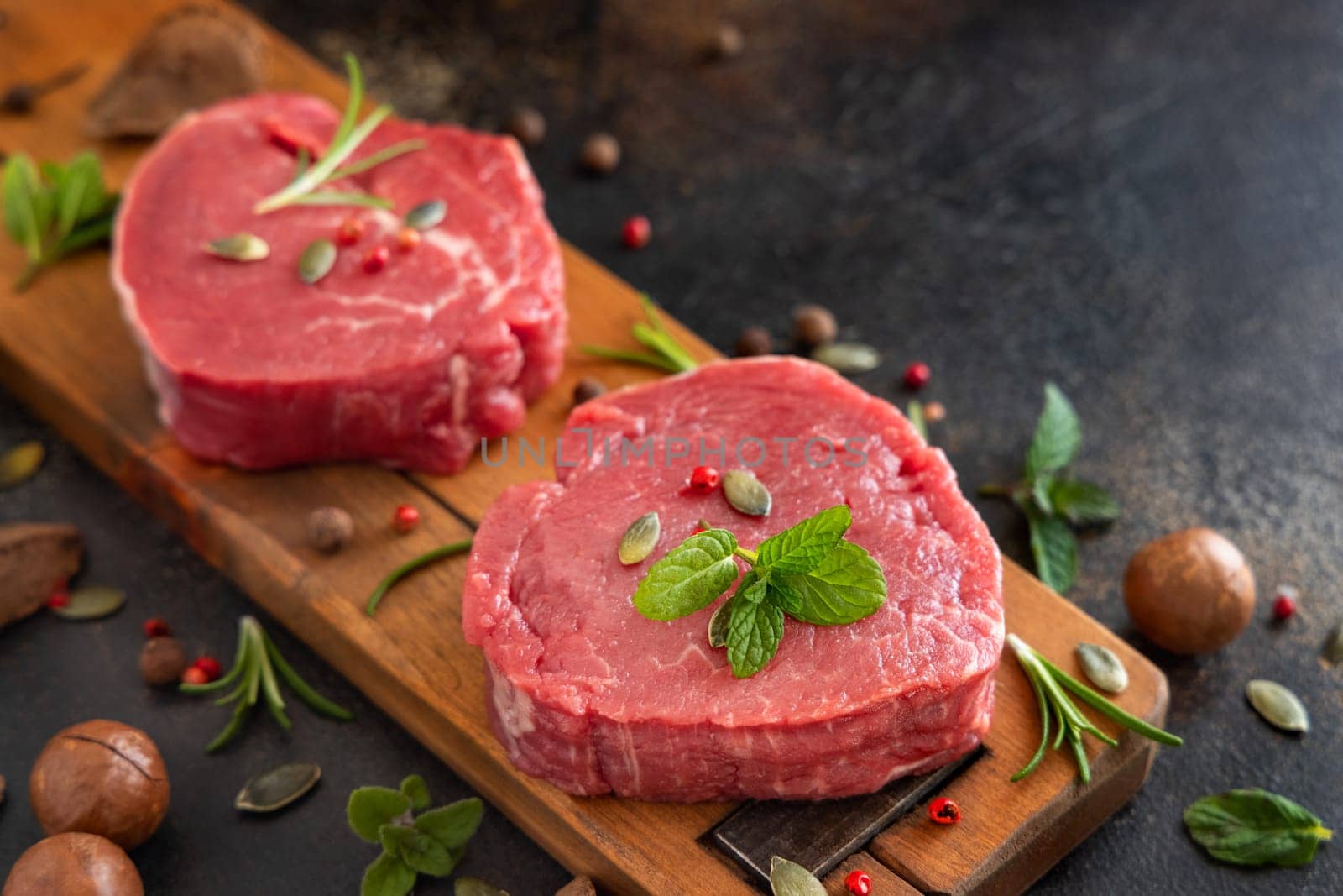 Raw beef fillet steaks on wooden board on dark table close up with copy space. Portioned filet mignons with spices and herbs. Concept of summer food, meat eaters.